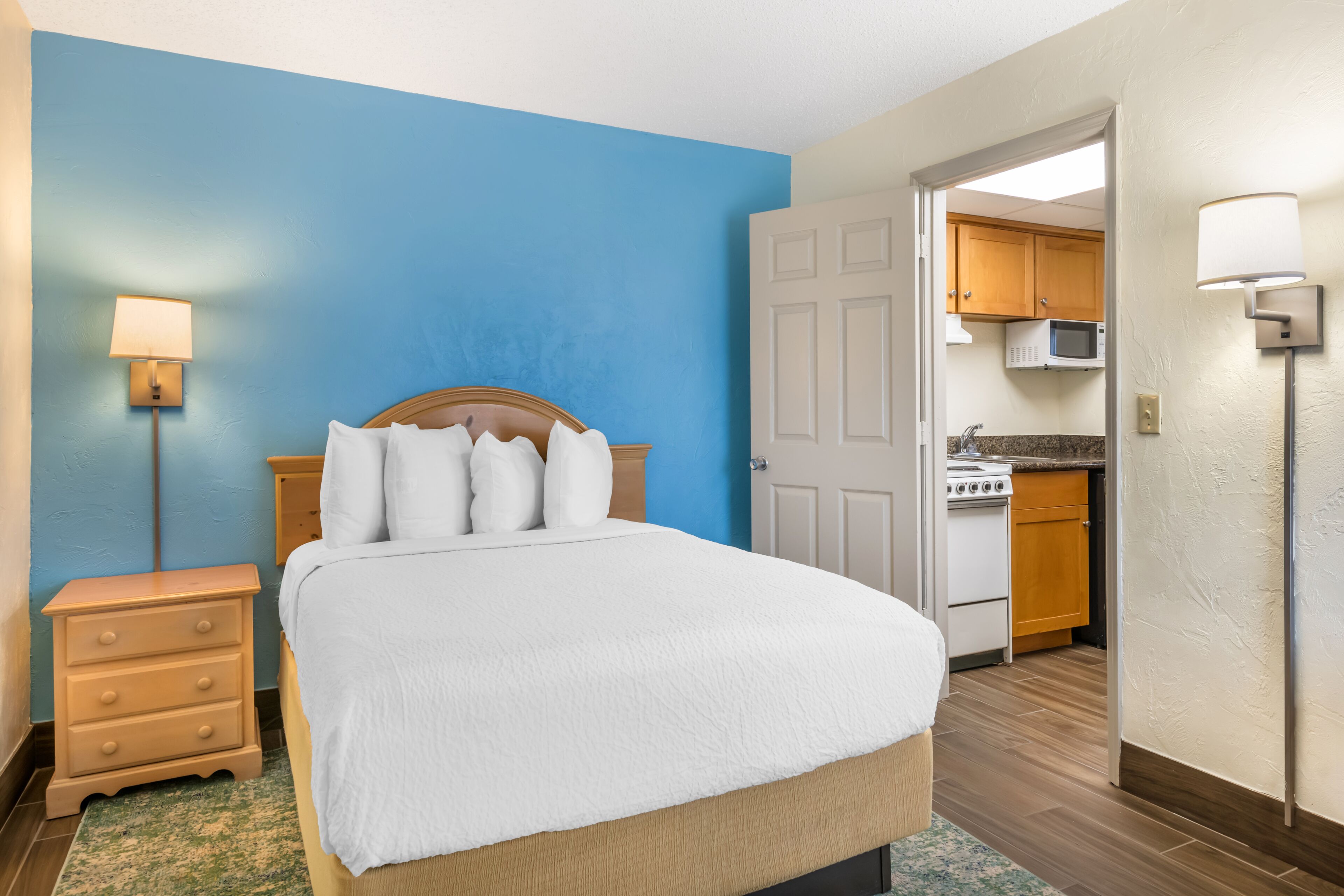 A bedroom with a bed, a nightstand, a lamp, and a view into a kitchen.