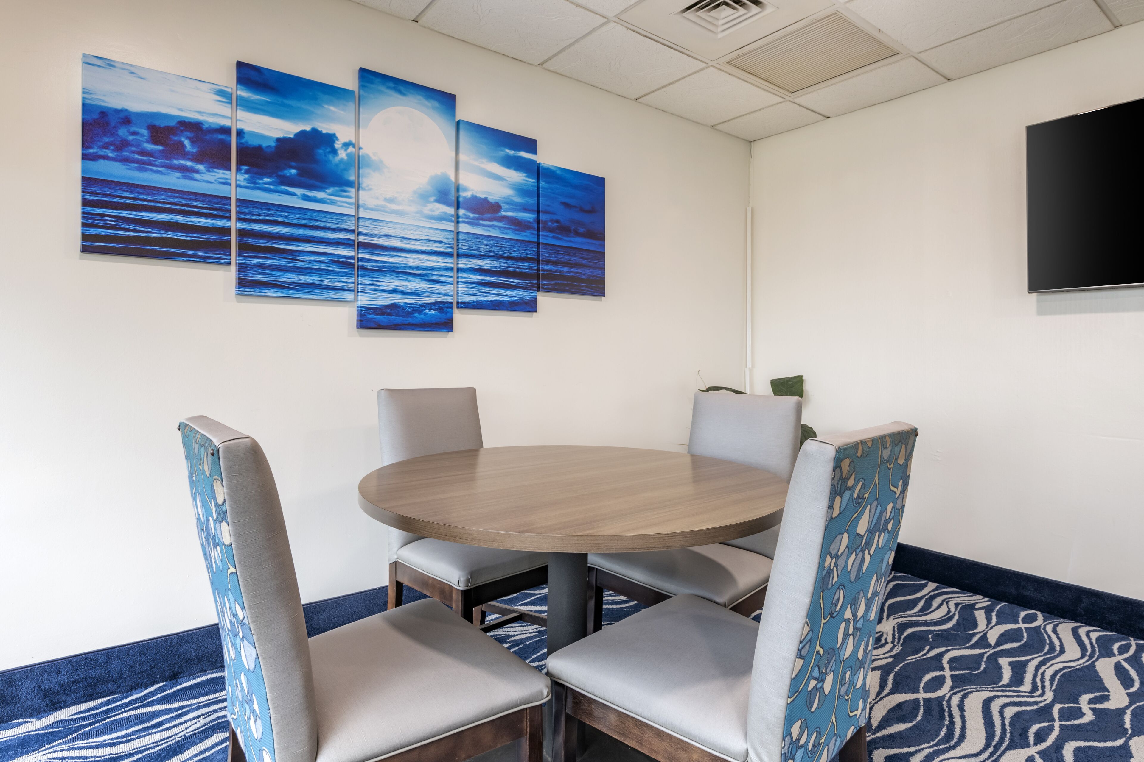 A dining area with a round table and six chairs, a large wall-mounted TV, and three framed artworks depicting a seascape.