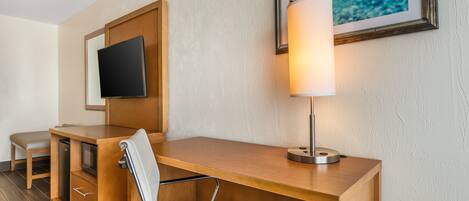 A hotel room with a wooden desk, a white chair, a flat-screen TV mounted on the wall, and a framed picture of a sea turtle.