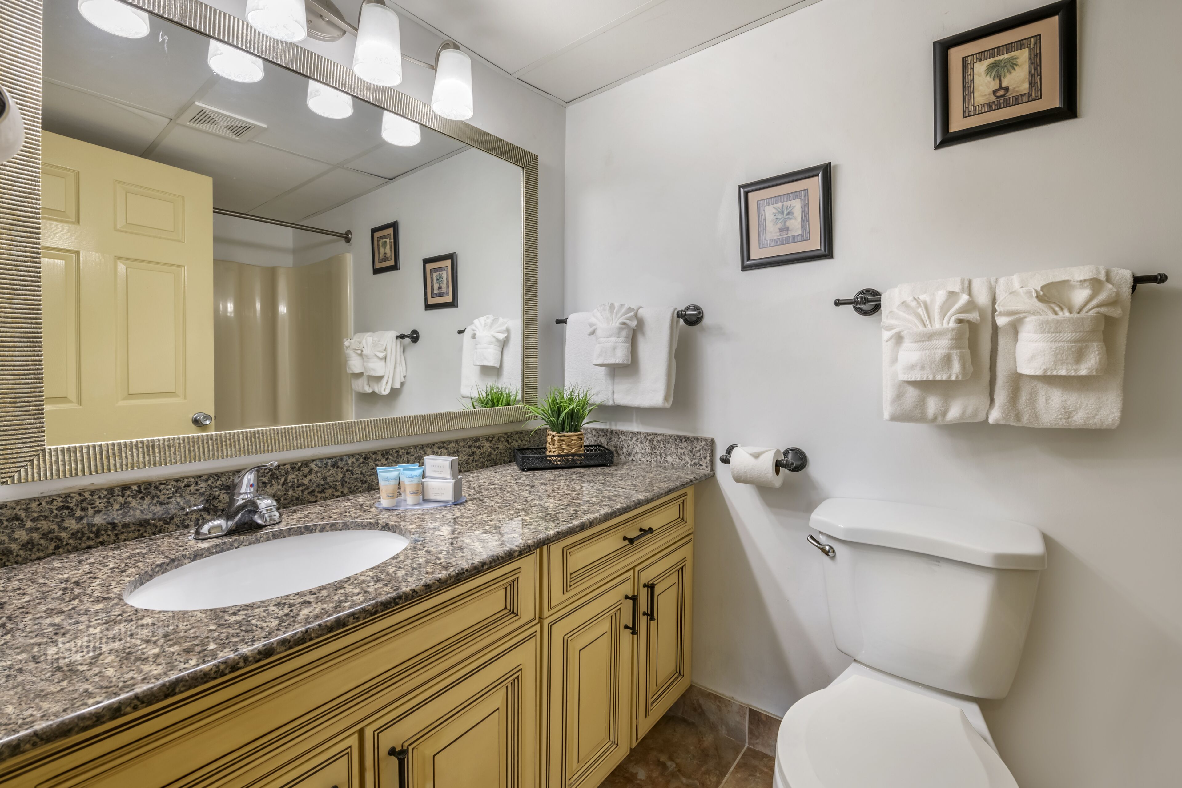 A bathroom with a granite countertop sink, a large mirror, a toilet, and framed pictures on the wall.