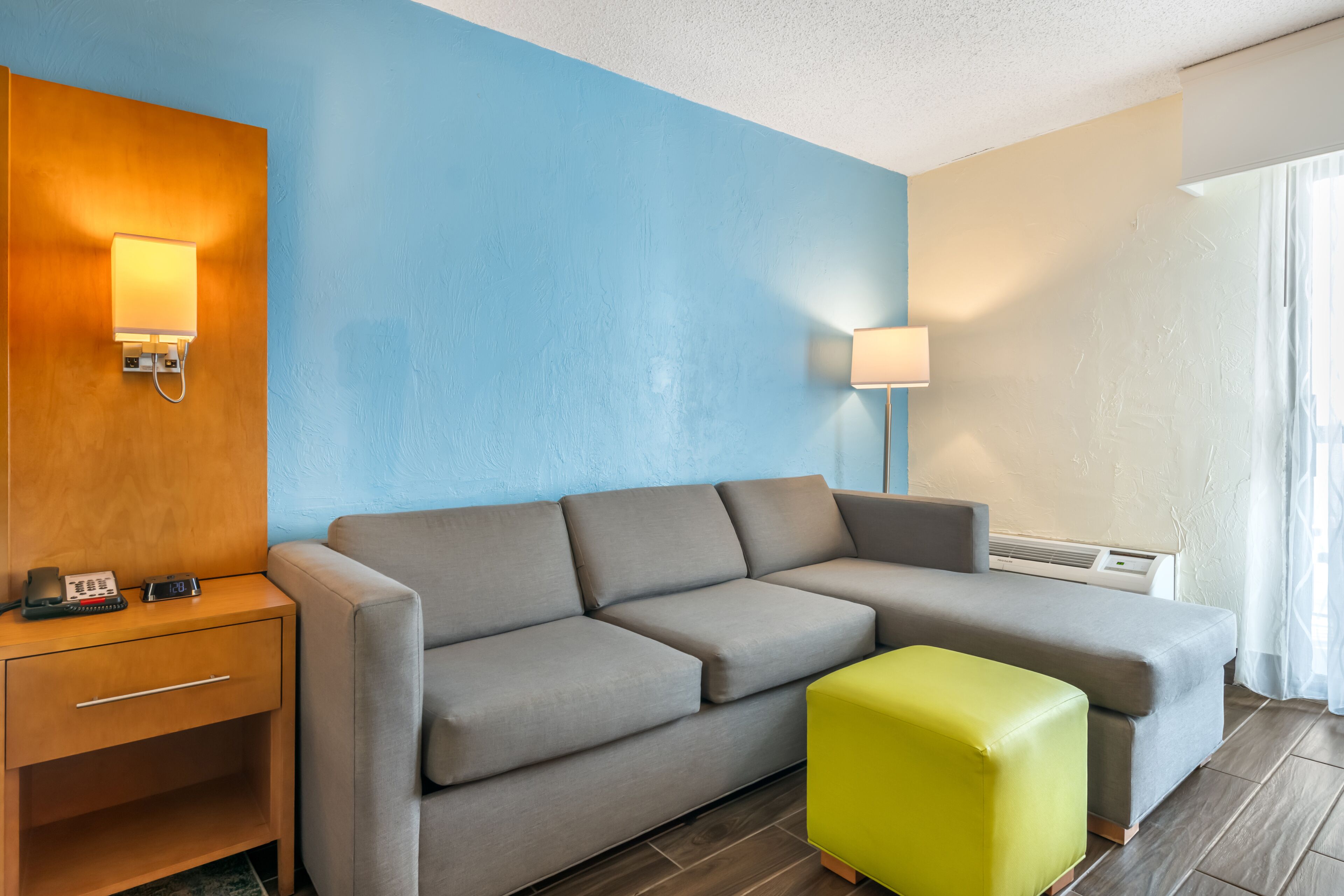 A modern hotel room with a grey sofa, a wooden nightstand with a phone, a bright green ottoman, and a blue wall.