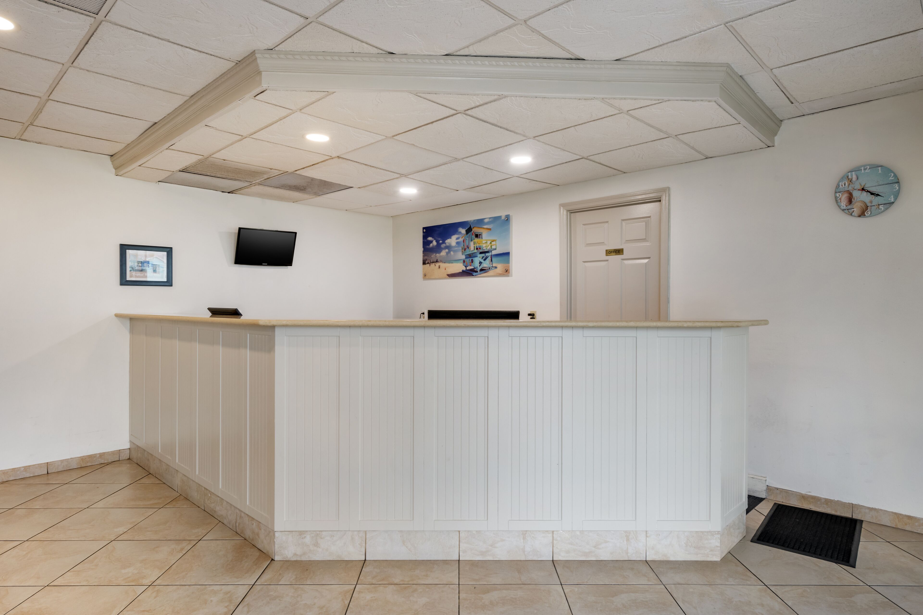 A reception area with a white reception desk, a flat-screen TV mounted on the wall, a painting of a beach scene, a clock, and a door.