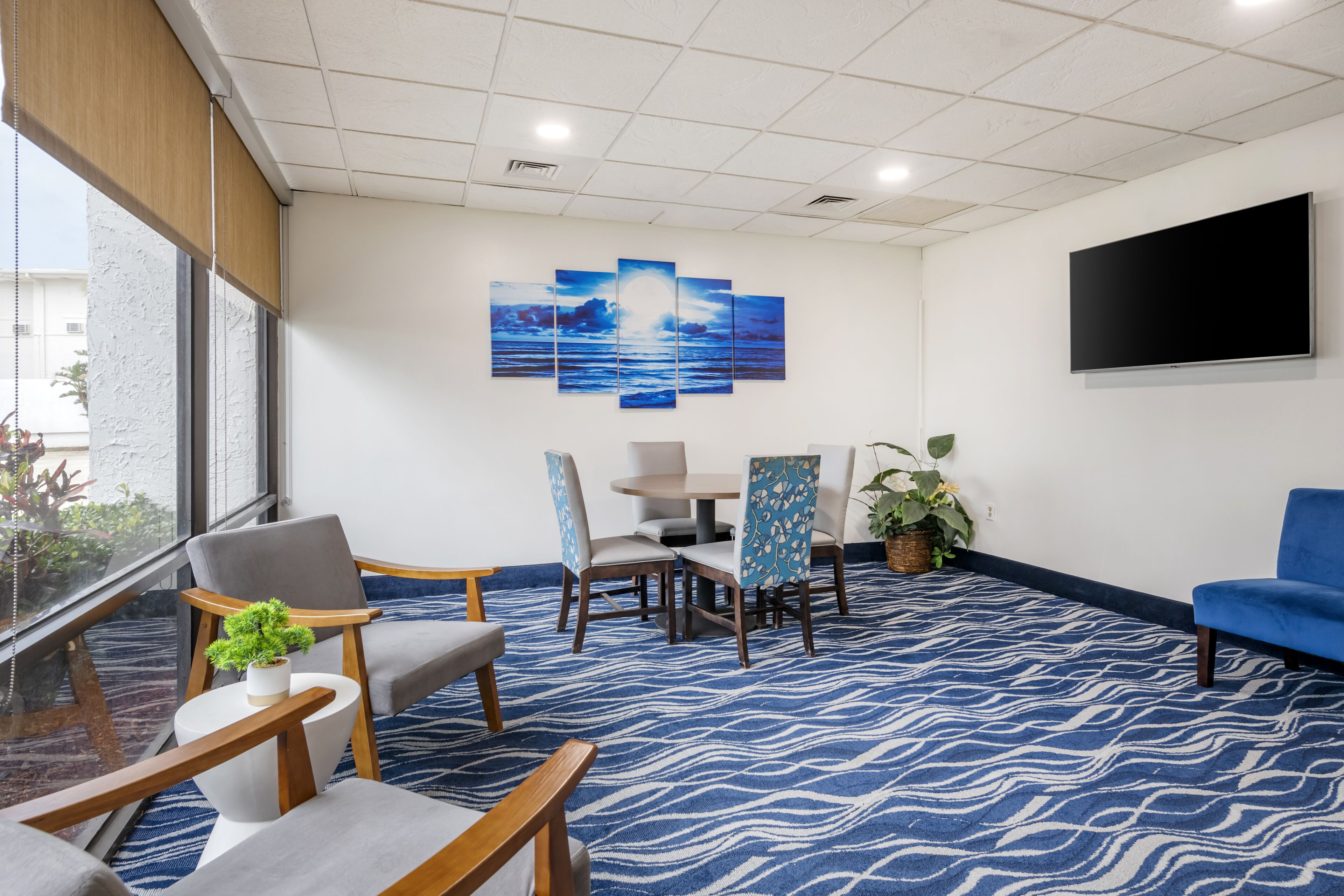Een hotel lobby met een blauw behangtapijt, een grote wandtelevisie en een zithoek met stoelen en een tafel.