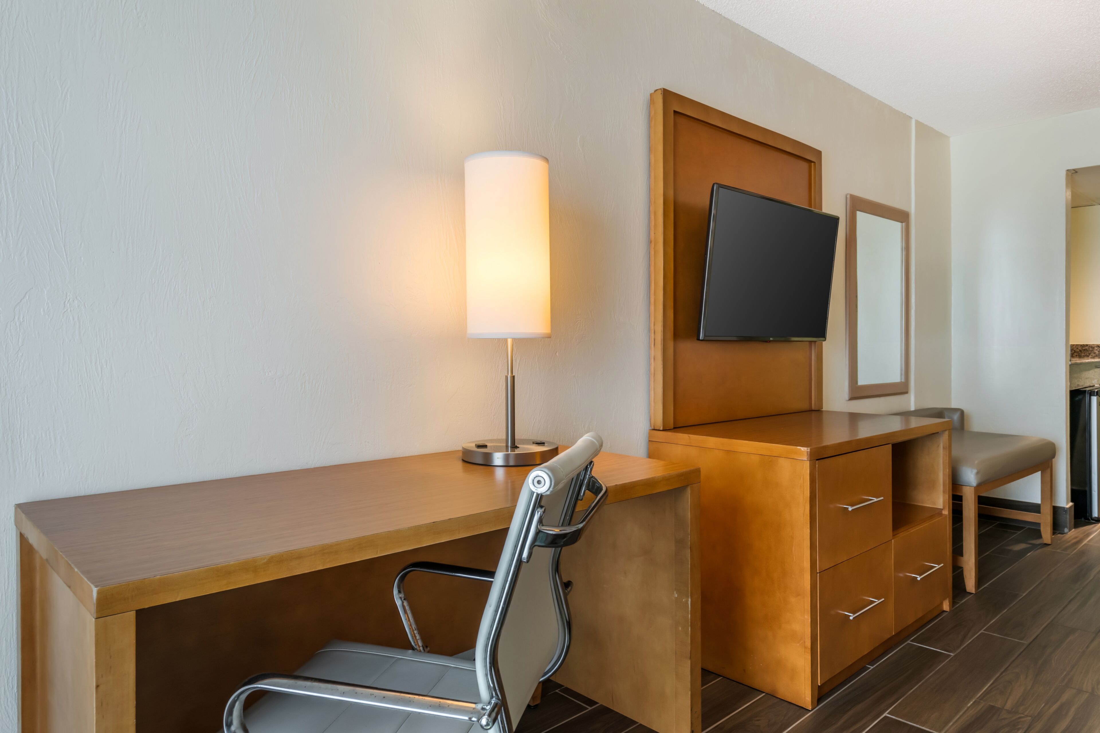 Una habitación de hotel con un escritorio de madera, una silla, un televisor montado en la pared, un espejo y una mesita de noche.