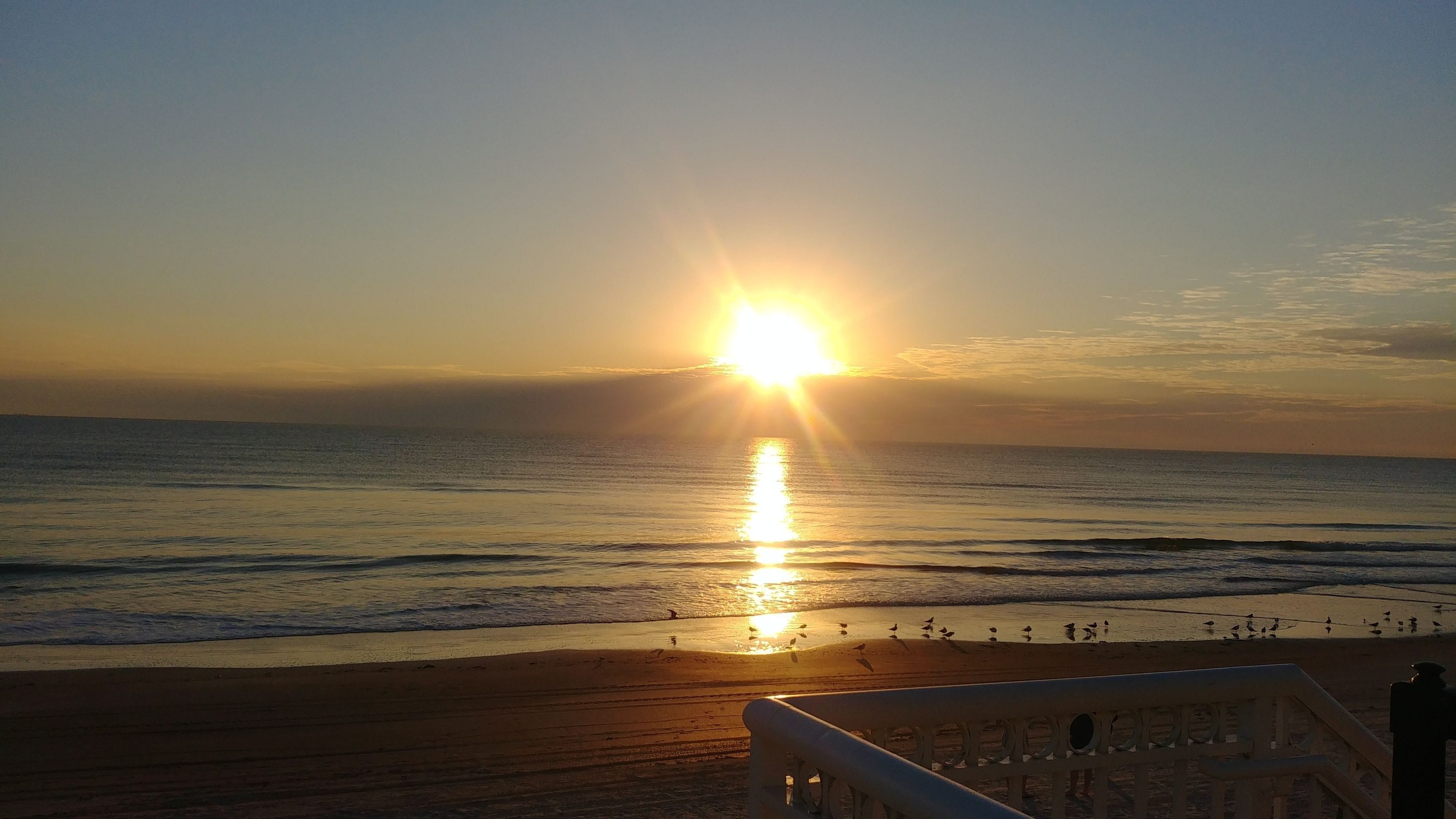 Une scène de plage au coucher du soleil sur l’océan, avec des vagues qui viennent doucement lécher le rivage et une rambarde en bois au premier plan.