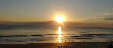 A beach scene with the sun setting over the ocean, waves gently lapping the shore, and a wooden railing in the foreground.