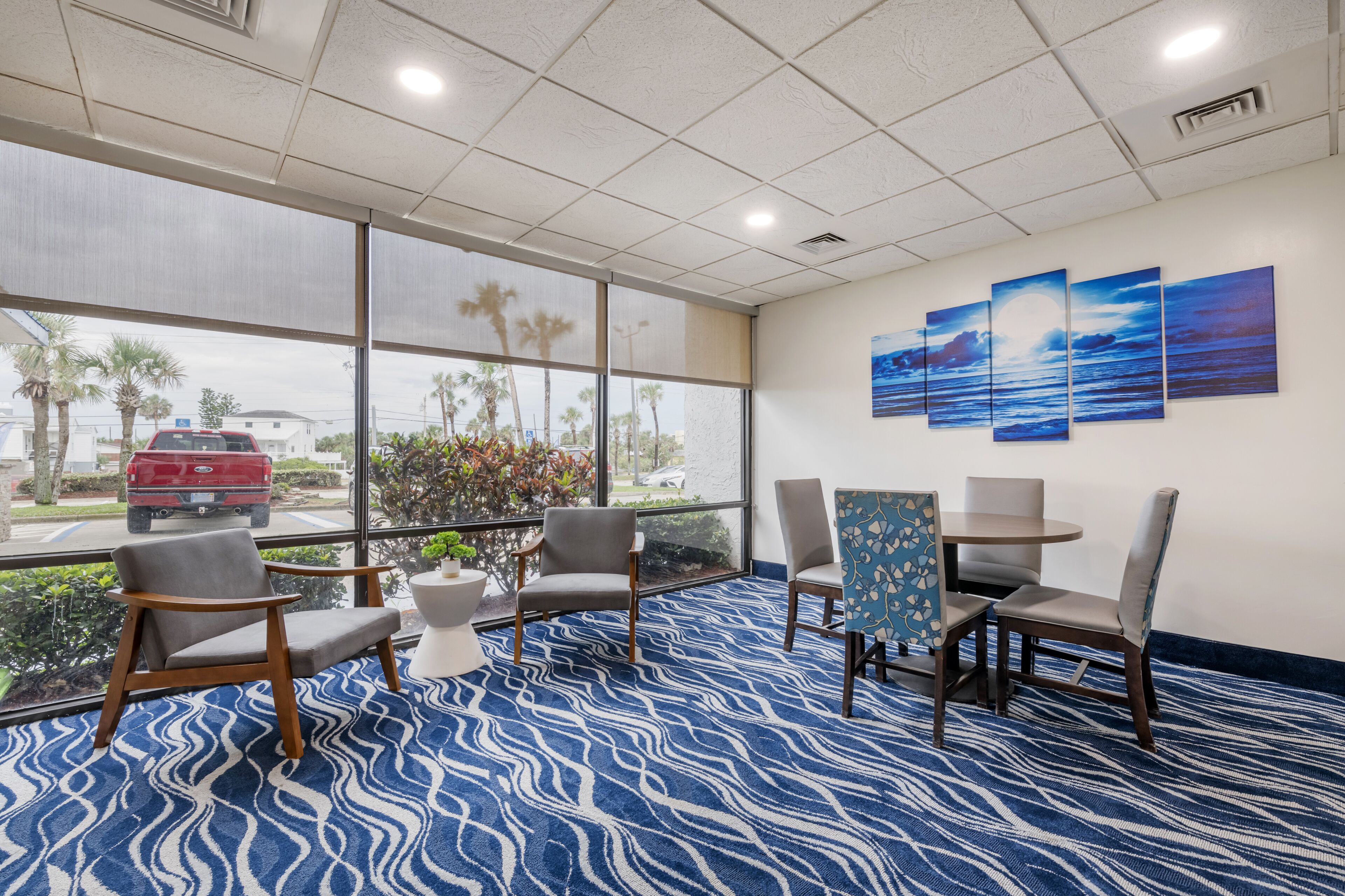 Un vestíbulo de hotel con alfombra de diseño azul, varias sillas, una pequeña mesa y grandes ventanales que ofrecen vistas a palmeras y un vehículo rojo en el exterior.