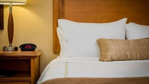 A hotel room with a wooden nightstand, a lamp, a digital clock, a bed with white bedding, and a woven throw pillow.