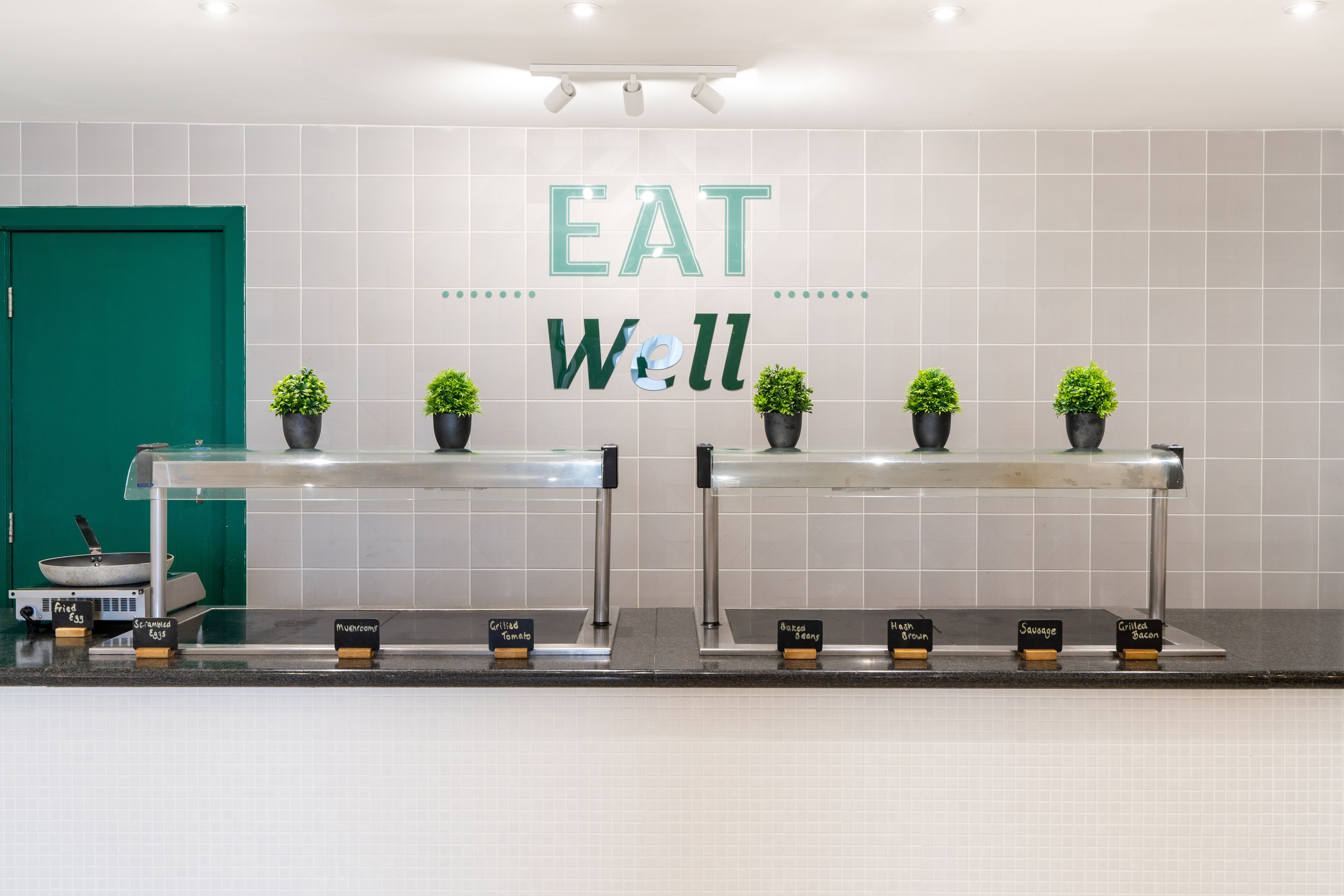 Eine Küchenarbeitsplatte mit einem Schild, auf dem 'EAT Well' steht, und Topfpflanzen. Beschriftungen zeigen verschiedene Lebensmittel wie gegrillte Tomaten und gebratenen Speck.