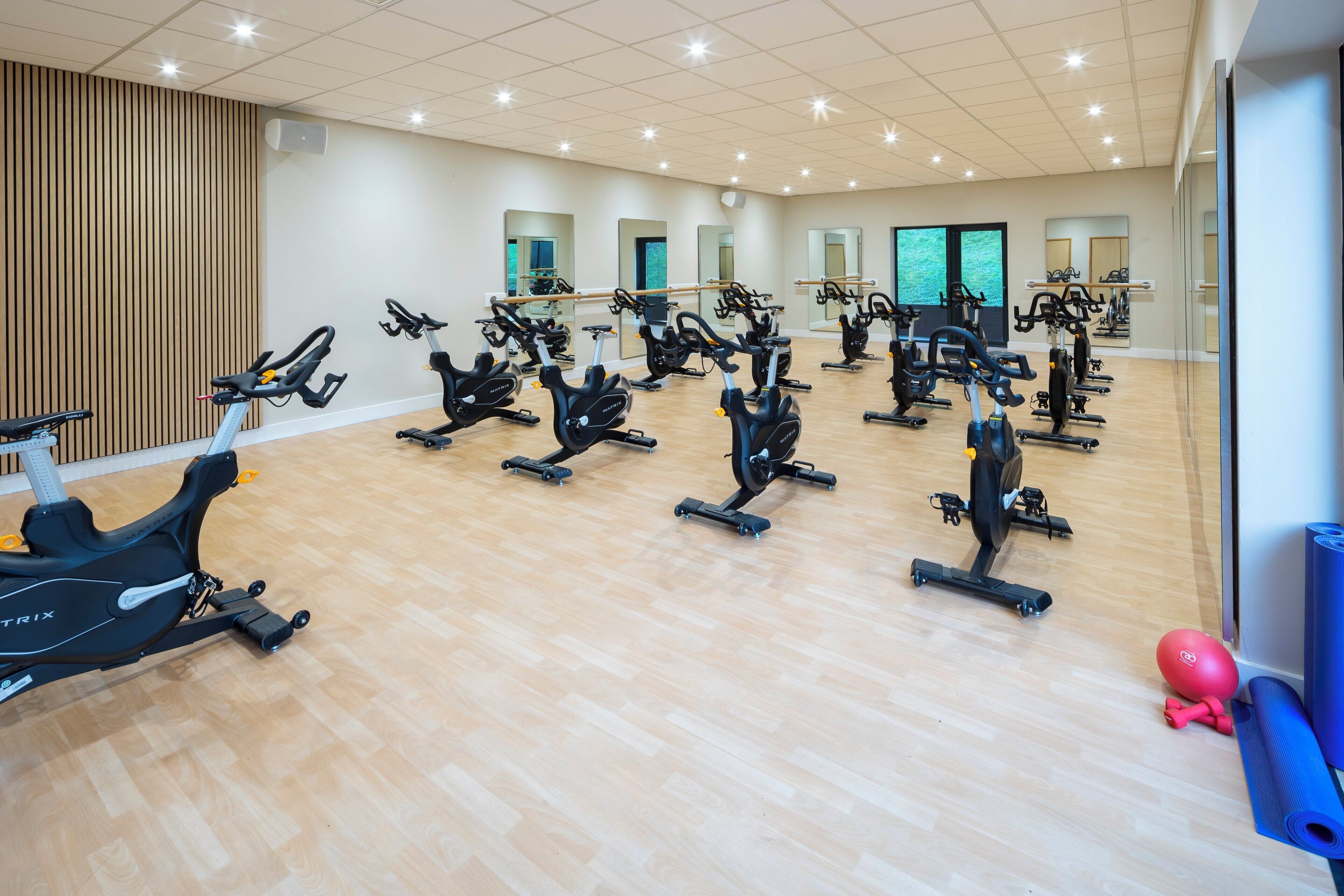Een ruime fitnessruimte met meerdere fietshallen en cardioapparaten, houten vloeren en grote spiegels.