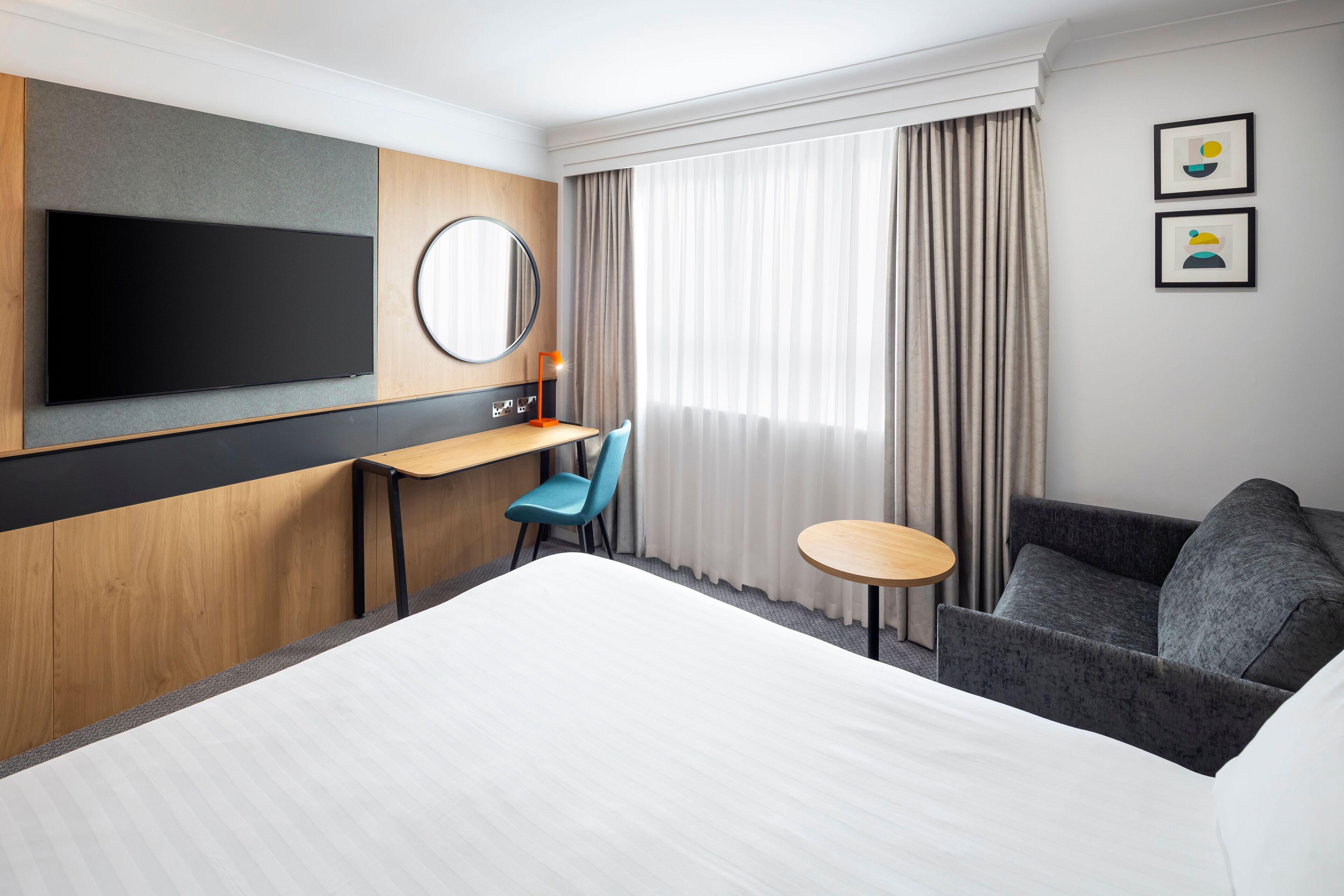 Een hotelkamer met een groot bed, een bureau met stoel, een klein rond tafeltje, een bank, een aan de muur bevestigde televisie, een spiegel en twee ingelijste foto's.