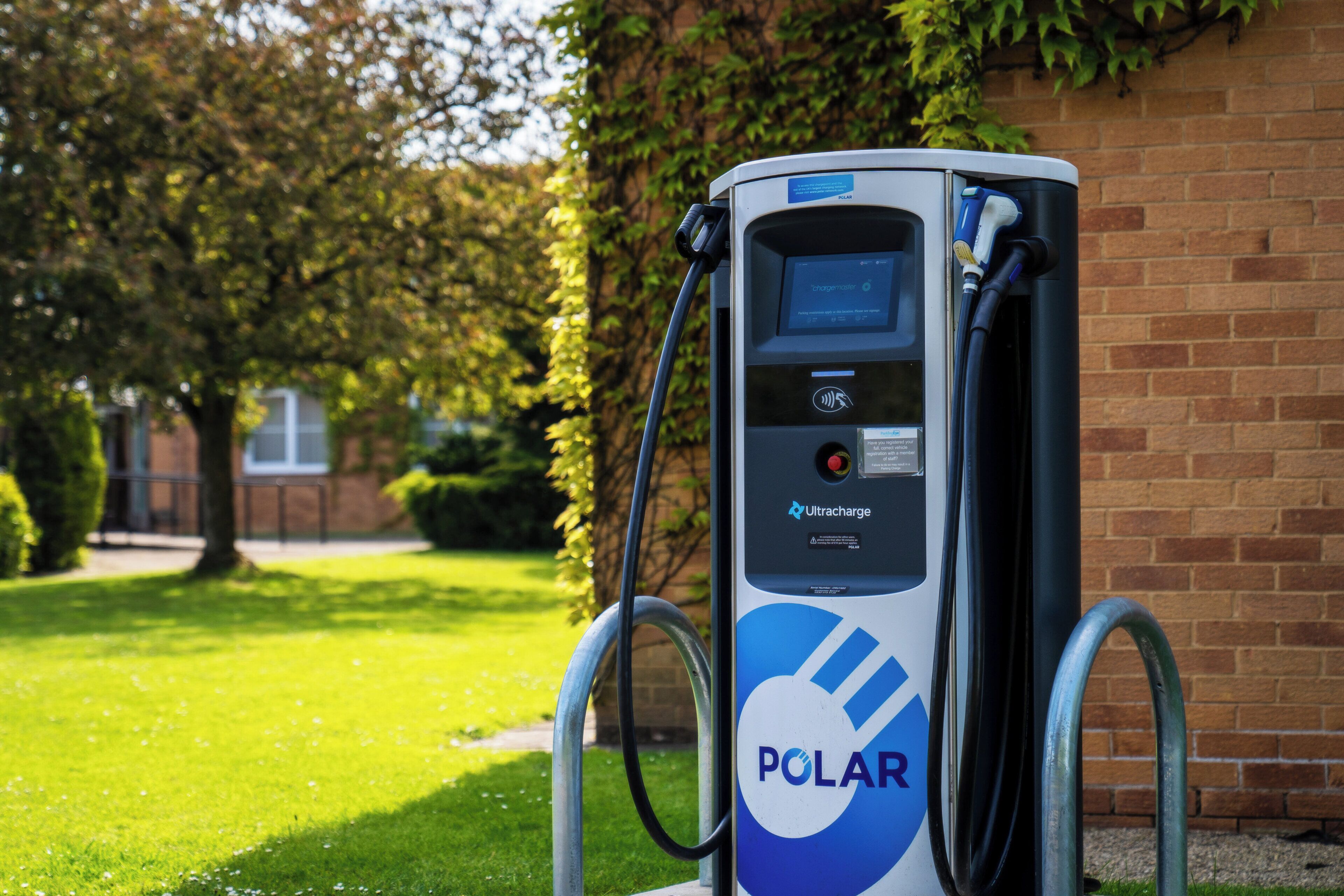 Une borne de recharge pour véhicules électriques POLAR, dotée d’un écran numérique, d’un lecteur de carte d’enregistrement et d’un bouton rouge, installée sur un mur de briques et entourée de verdure.