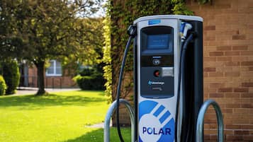 A POLAR electric vehicle charging station with a digital display, a registration card slot, and a red button, set against a brick wall and greenery.