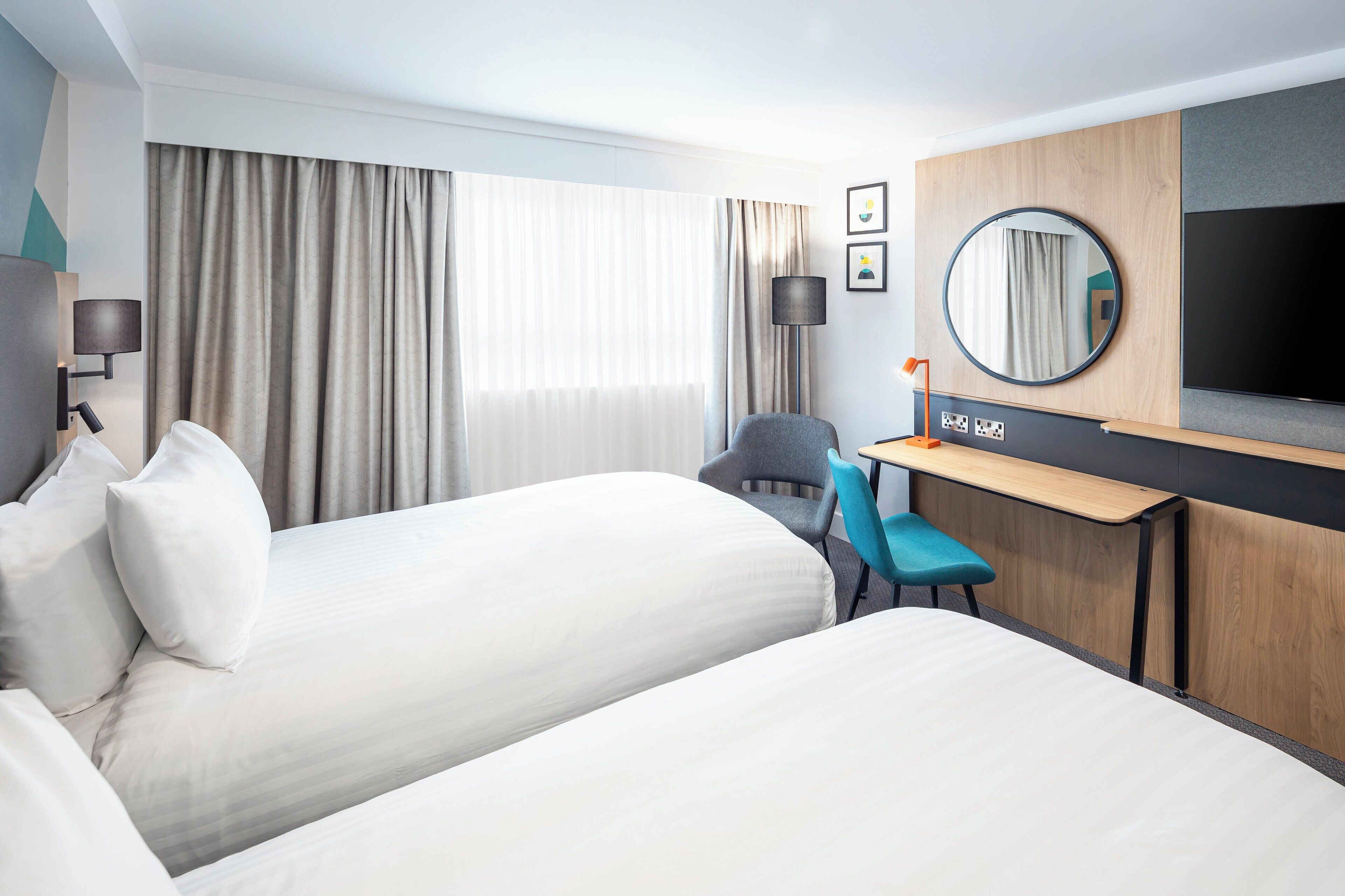 A hotel room with two beds, a desk, a chair, a TV, and a large window with curtains.