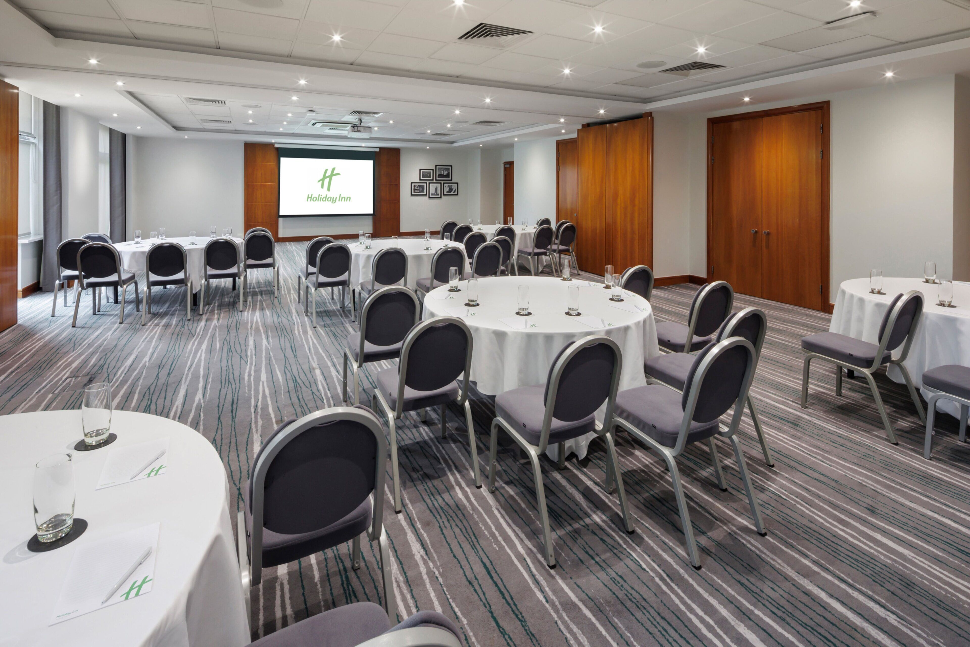 Un salón de conferencias con mesas y sillas redondas, una pantalla de proyector mostrando 'Holiday Inn', y piso alfombrado.