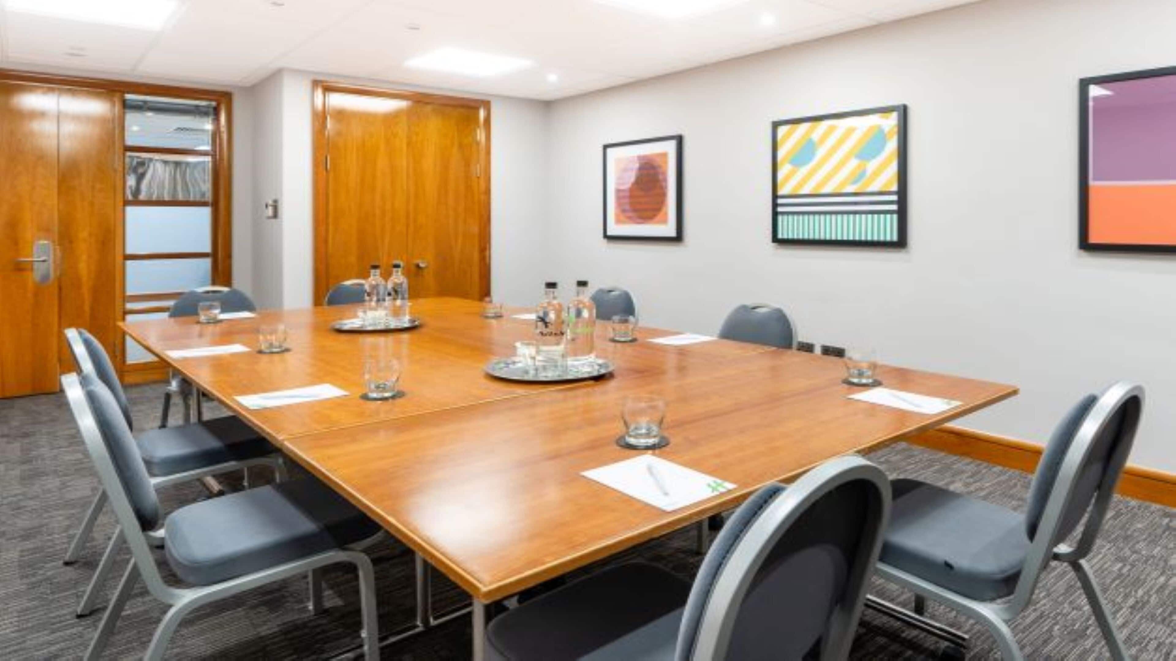 A conference room with a wooden table, grey chairs, and framed artwork on the walls.