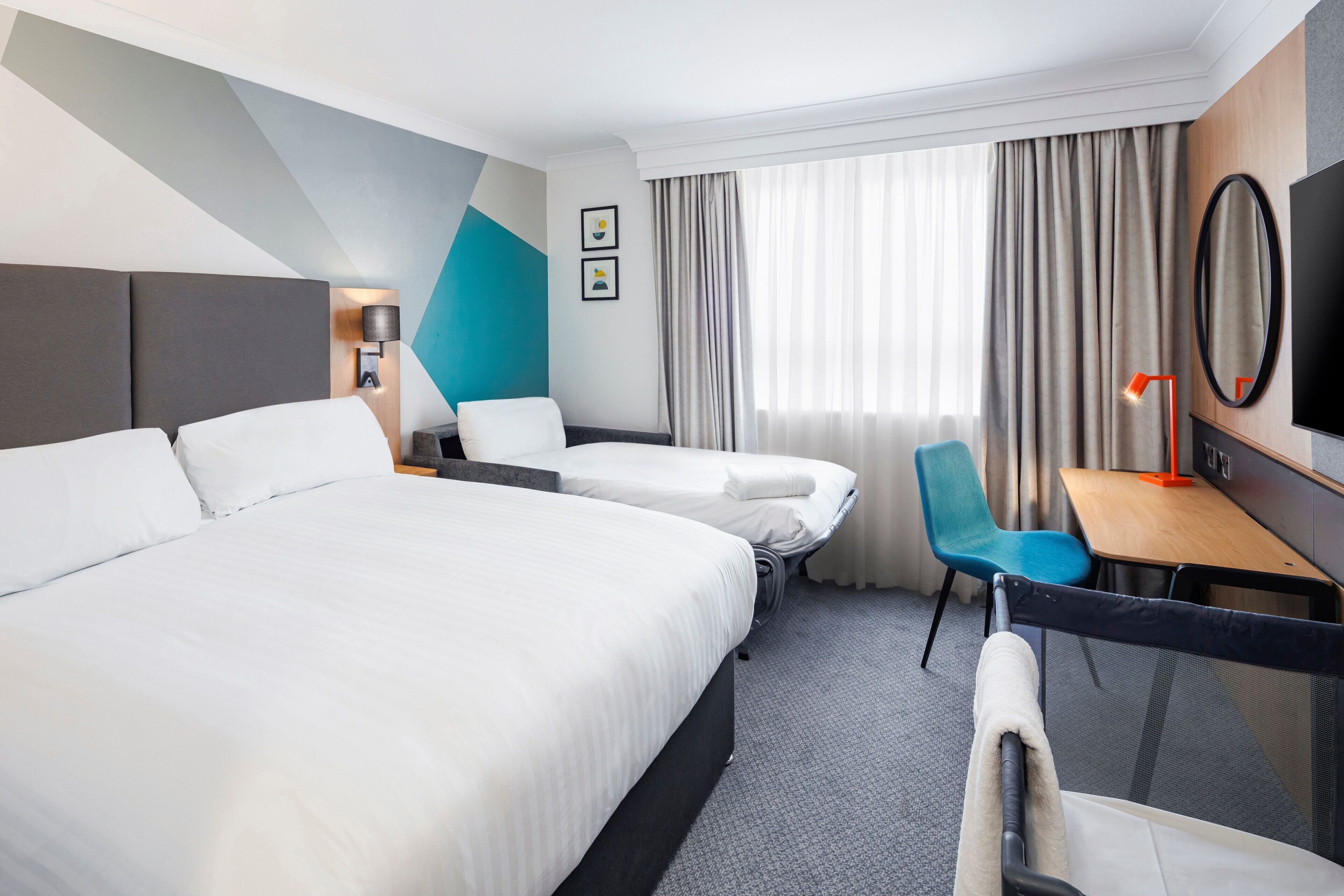 A hotel room with two beds, a desk, a chair, a TV, and a window with curtains.
