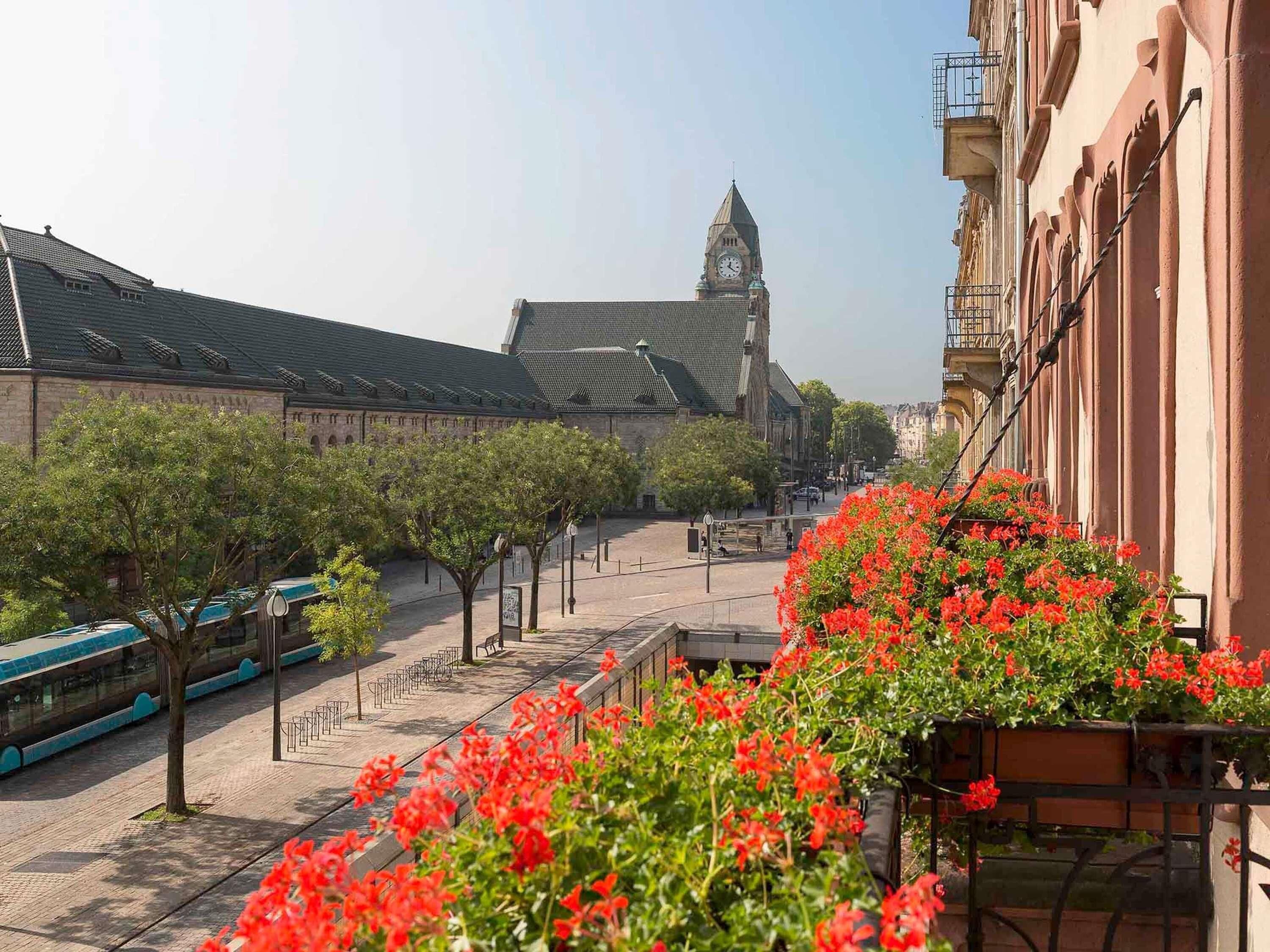 Vista da rua com um edifício com torre do relógio, árvores e um passeio decorado com flores.