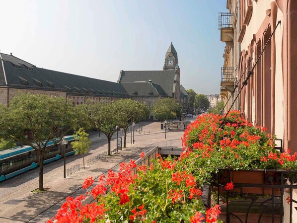 Vista de la calle con un edificio que tiene una torre del reloj, árboles y un paseo bordeado de flores.
