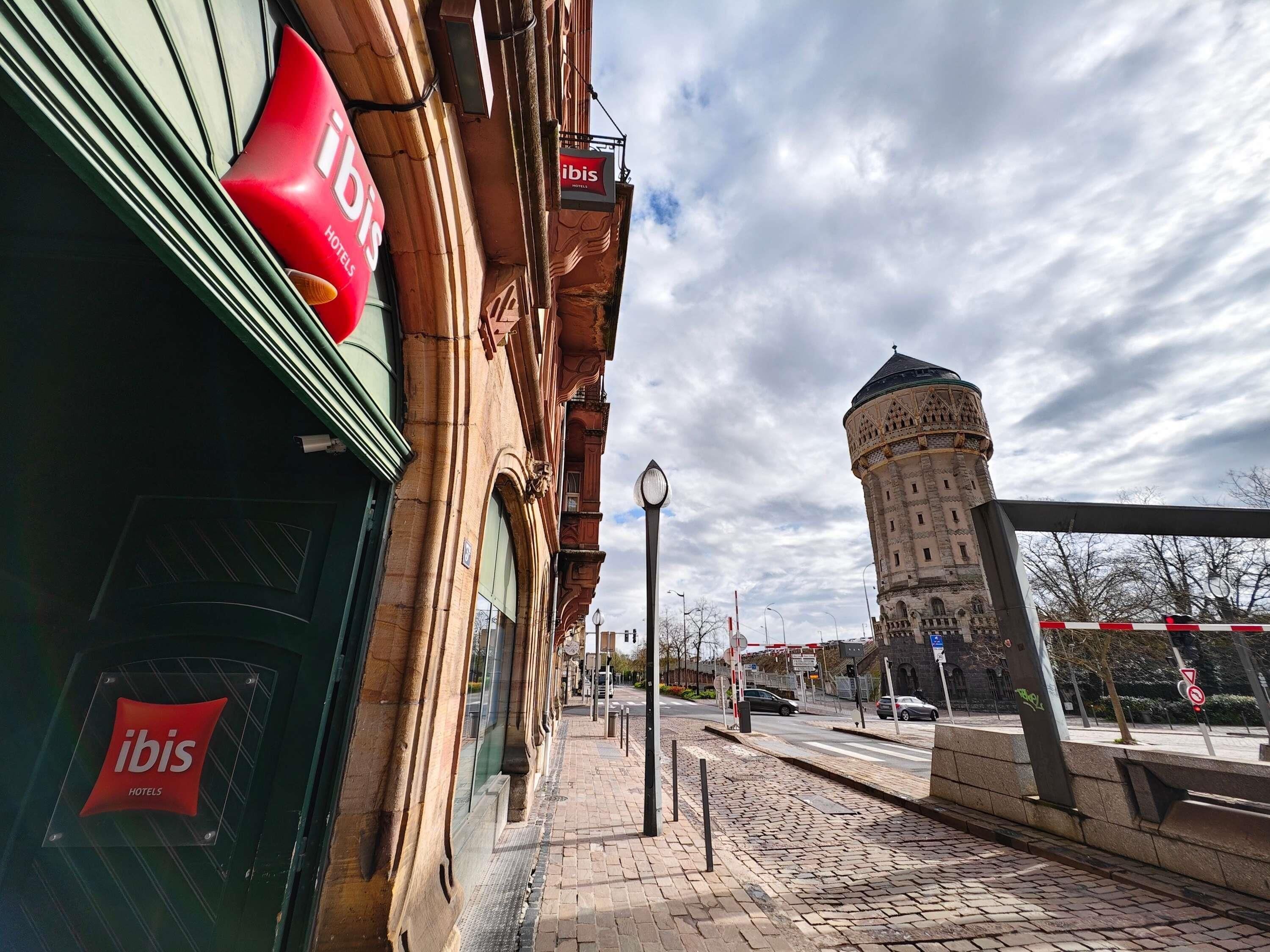 A historic building with a prominent tower, a red ibis hotel sign, and a cobblestone street.