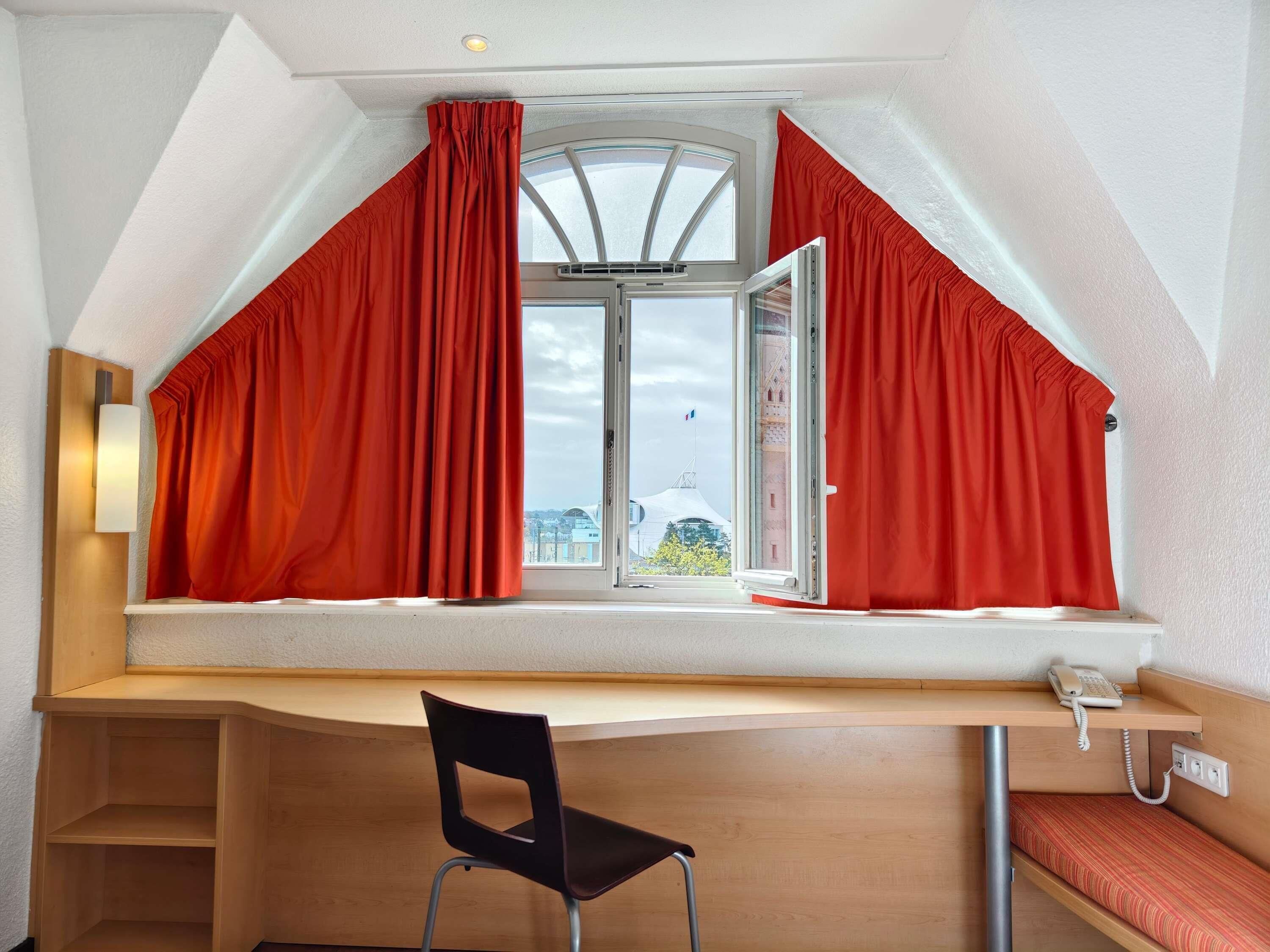 Una habitación con techo inclinado, un escritorio con silla, un teléfono y una ventana con cortinas rojas.