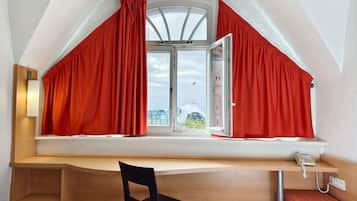 Una habitación con techo inclinado, un escritorio con silla, un teléfono y una ventana con cortinas rojas.