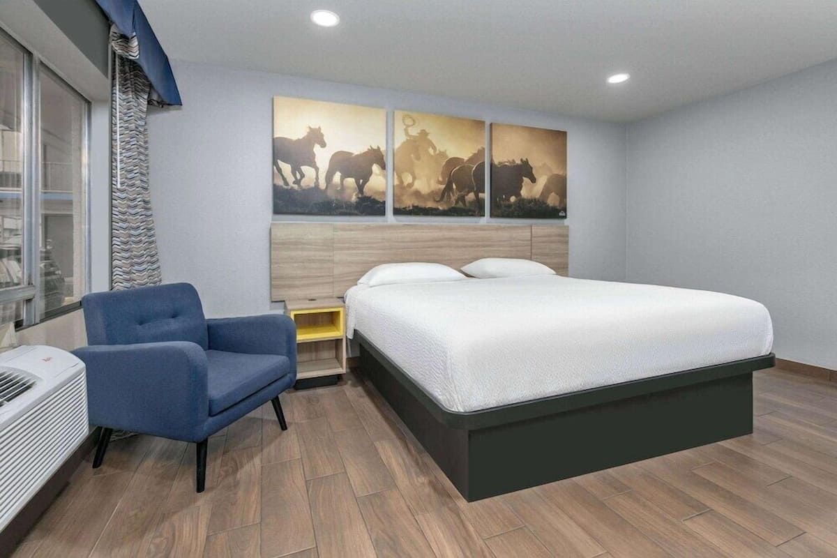 A hotel room with a bed, a chair, a nightstand, a wall-mounted air conditioner, and a window with a curtain.