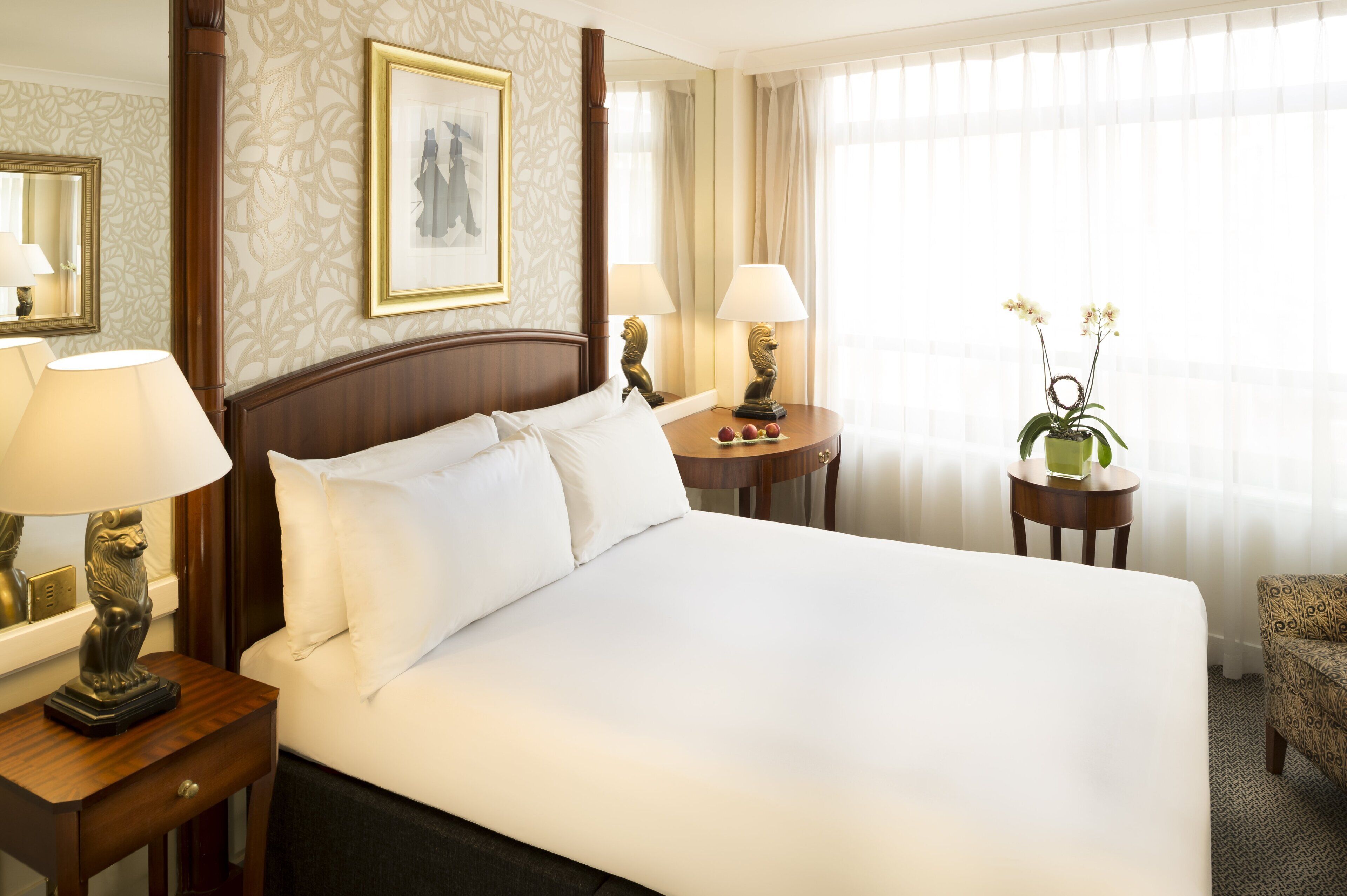 A hotel room with a large bed, two bedside tables with lamps, a small round table with a vase of flowers, and a chair.