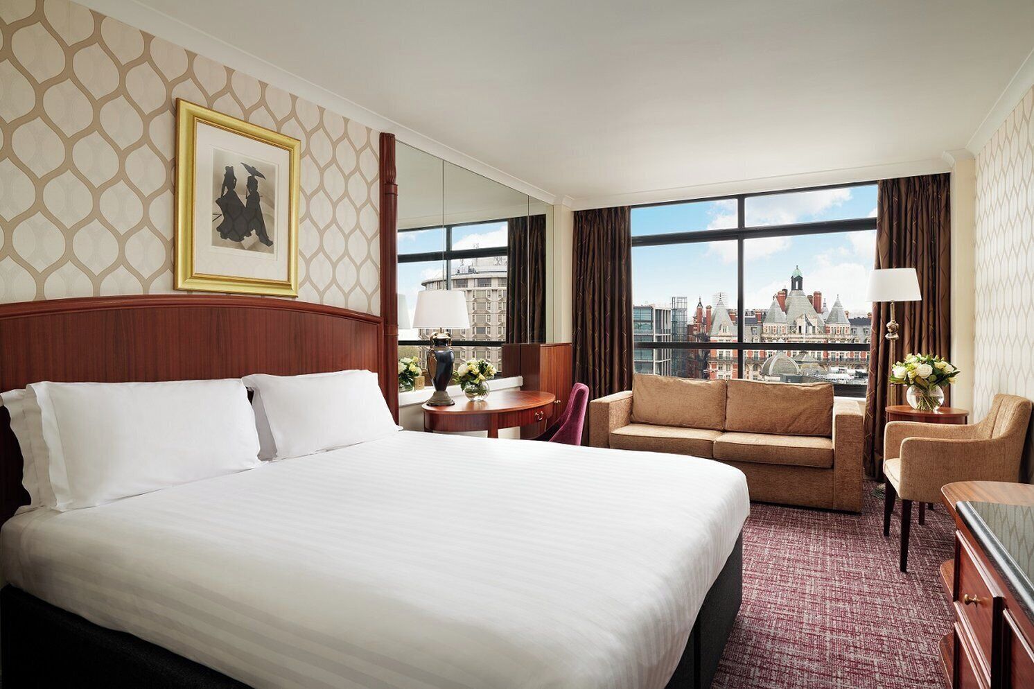 A hotel room with a large bed, a sofa, a desk, and a chair. There is a window with a city view.