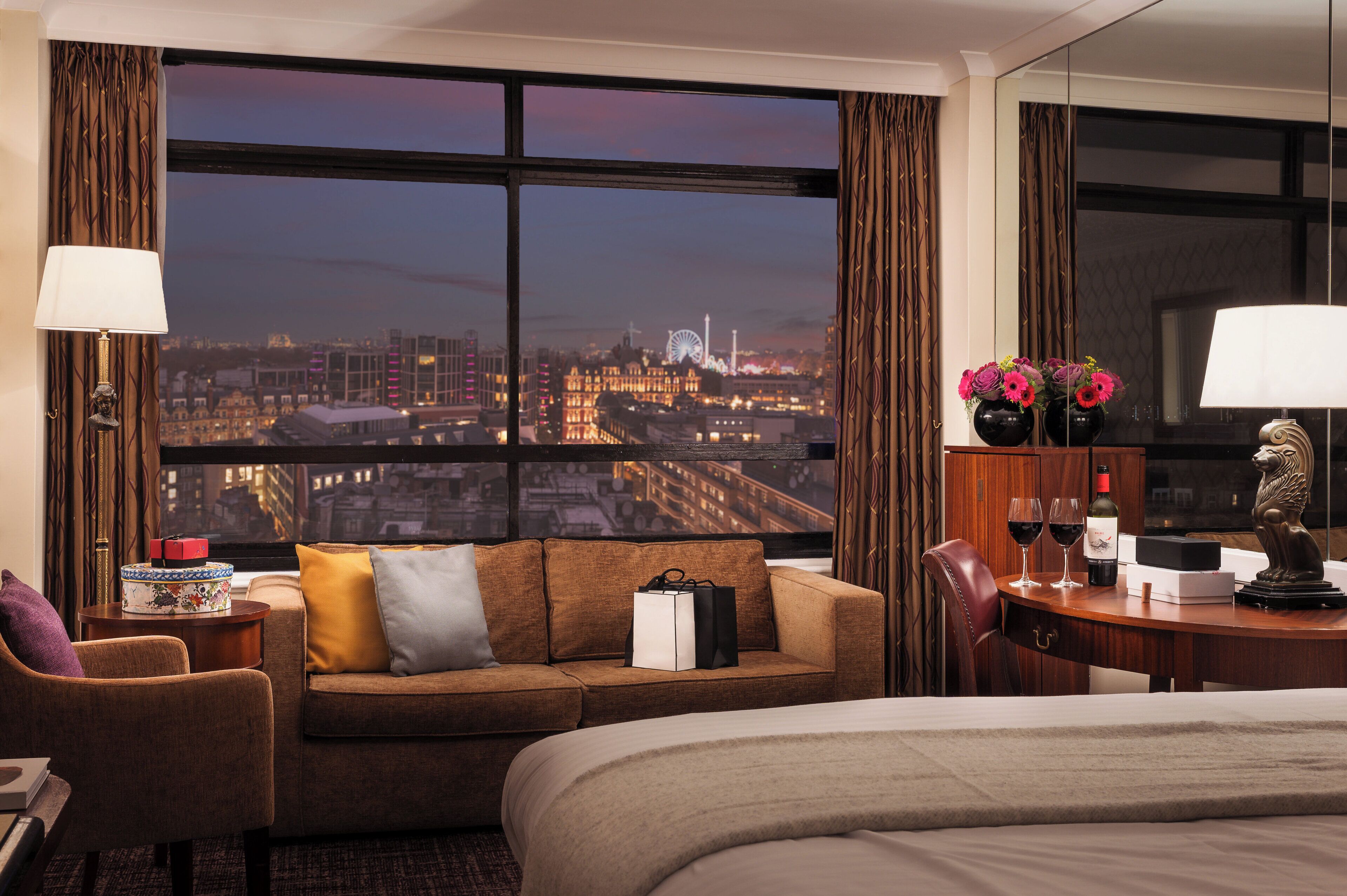 A hotel room with a large window offering a city view, a sofa, a desk with a lamp, and a bed.
