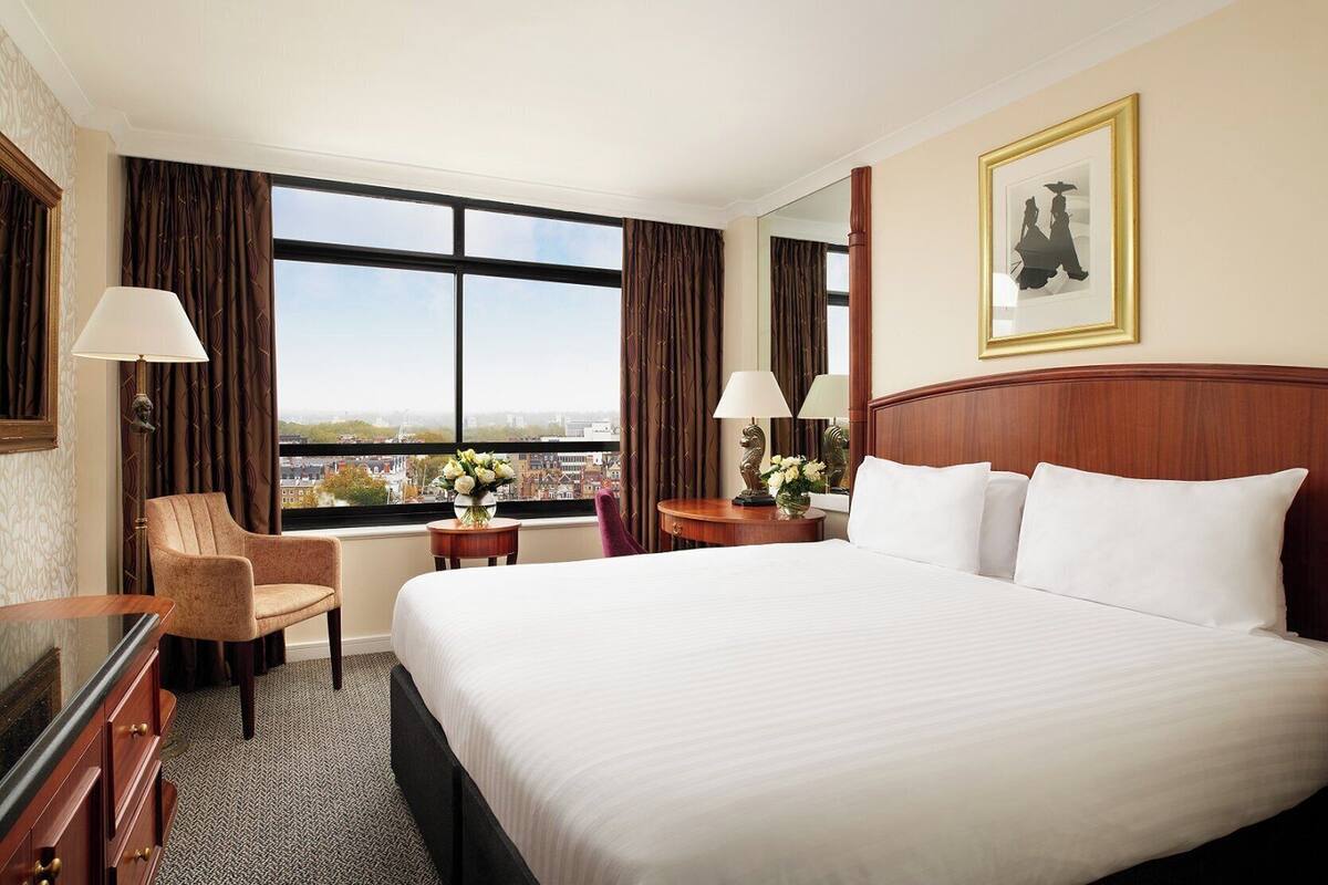 A hotel room with a large bed, a desk, a chair, a window with a city view, and a framed picture on the wall.