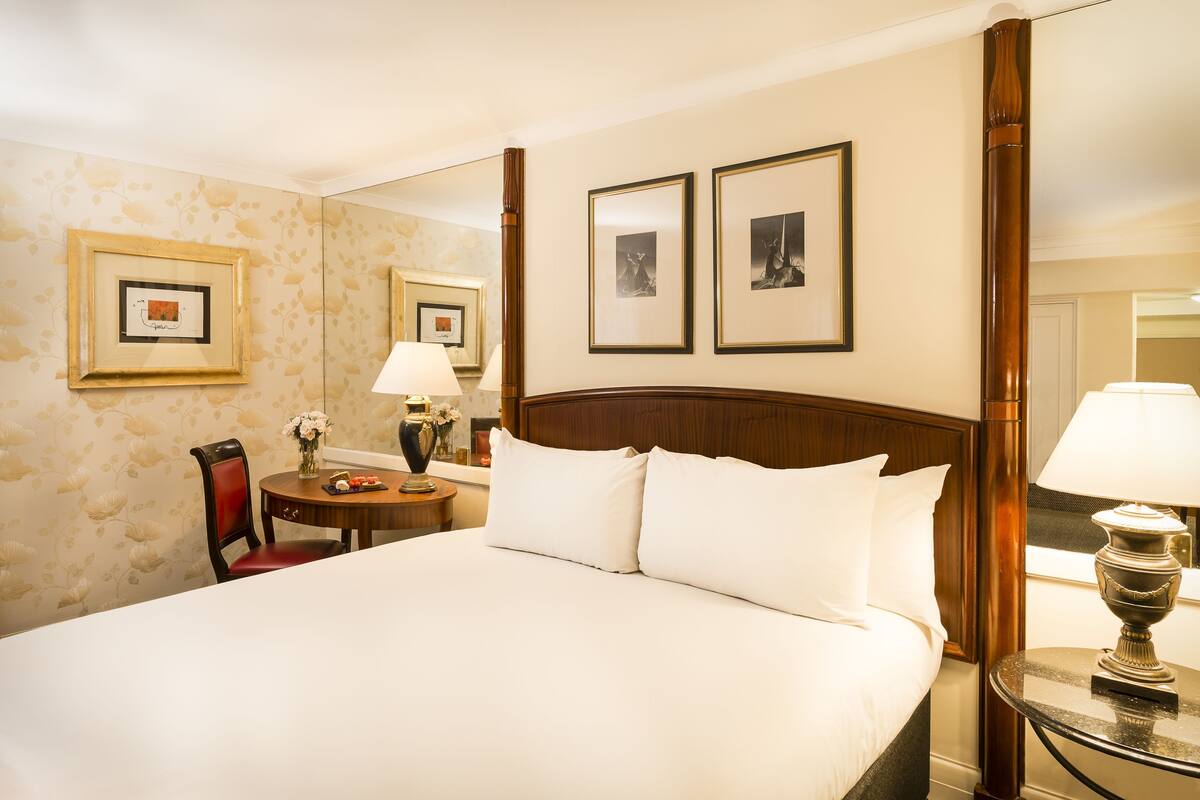 A hotel room with a large bed, a round table with a lamp, a chair, and framed pictures on the wall.