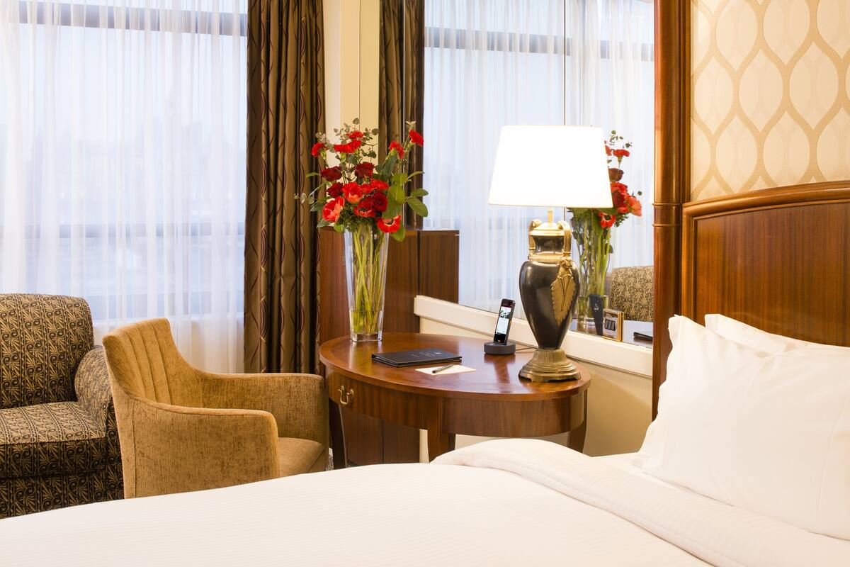 A hotel room with a bed, a desk with a lamp, a vase with flowers, a chair, and a view through the window.