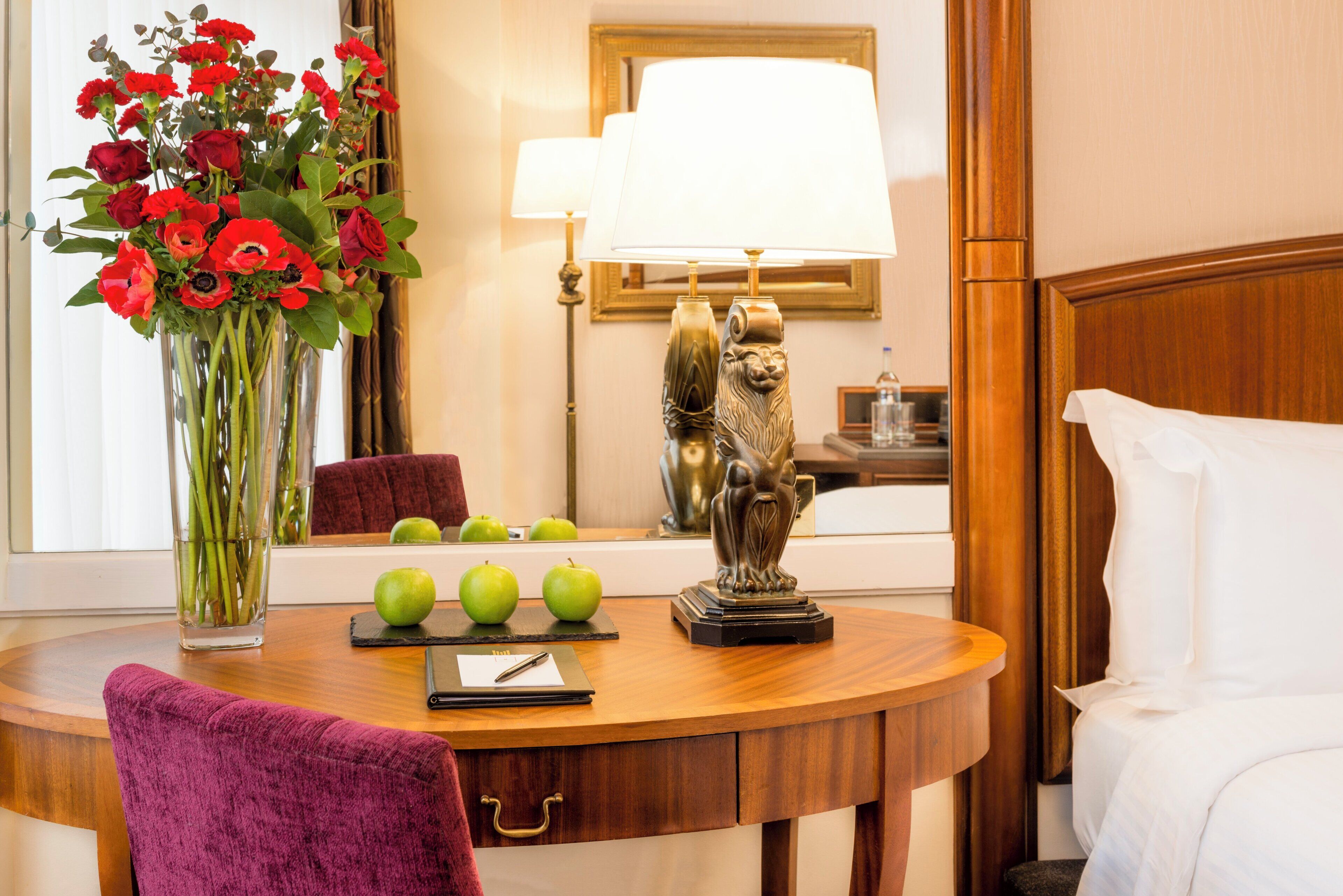 A hotel room with a wooden desk, a vase of red flowers, a lamp, and a bed with white linens.
