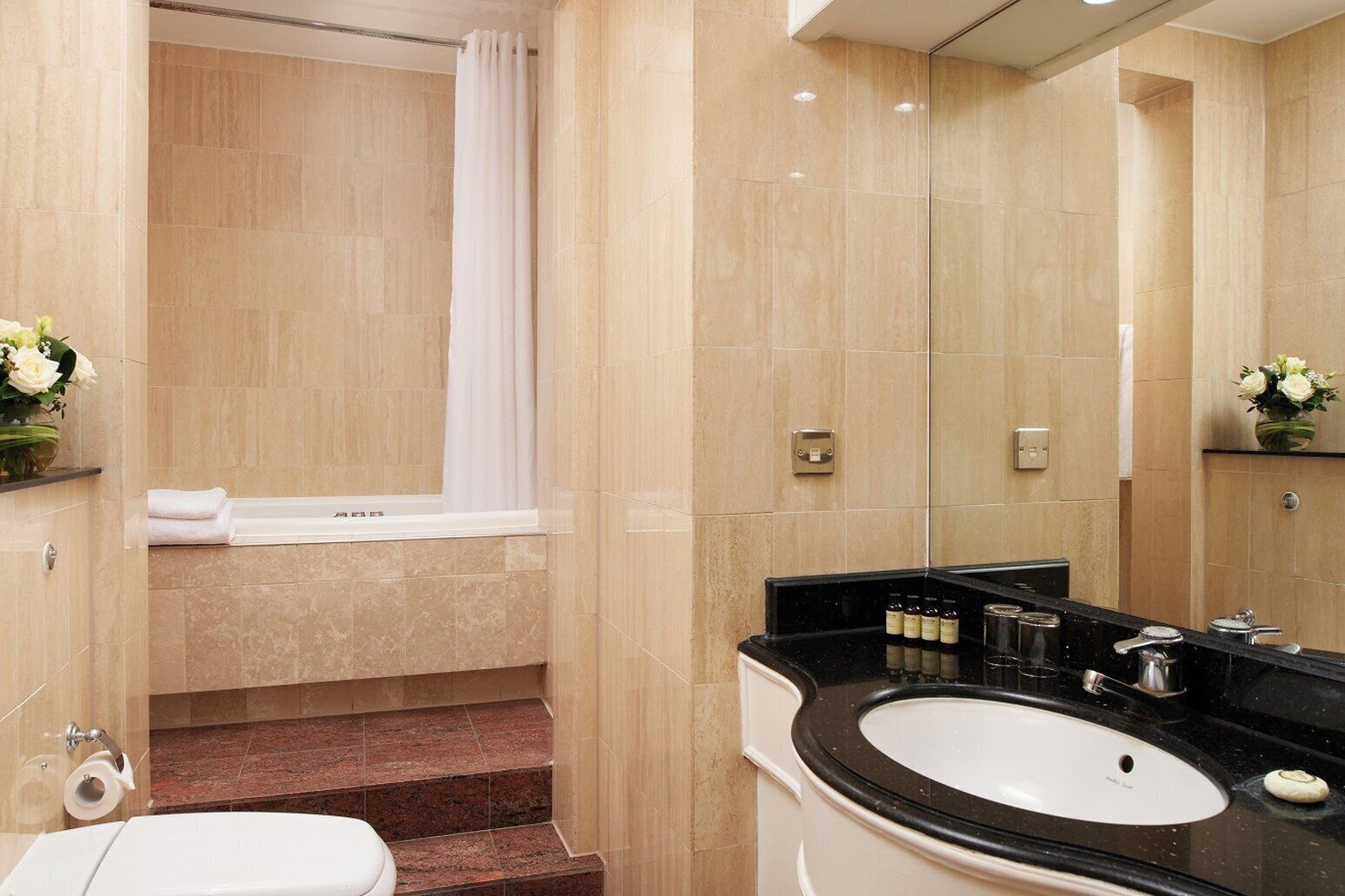 A modern bathroom with a bathtub, a sink, and a toilet. There are beige tiles on the walls and a black countertop.
