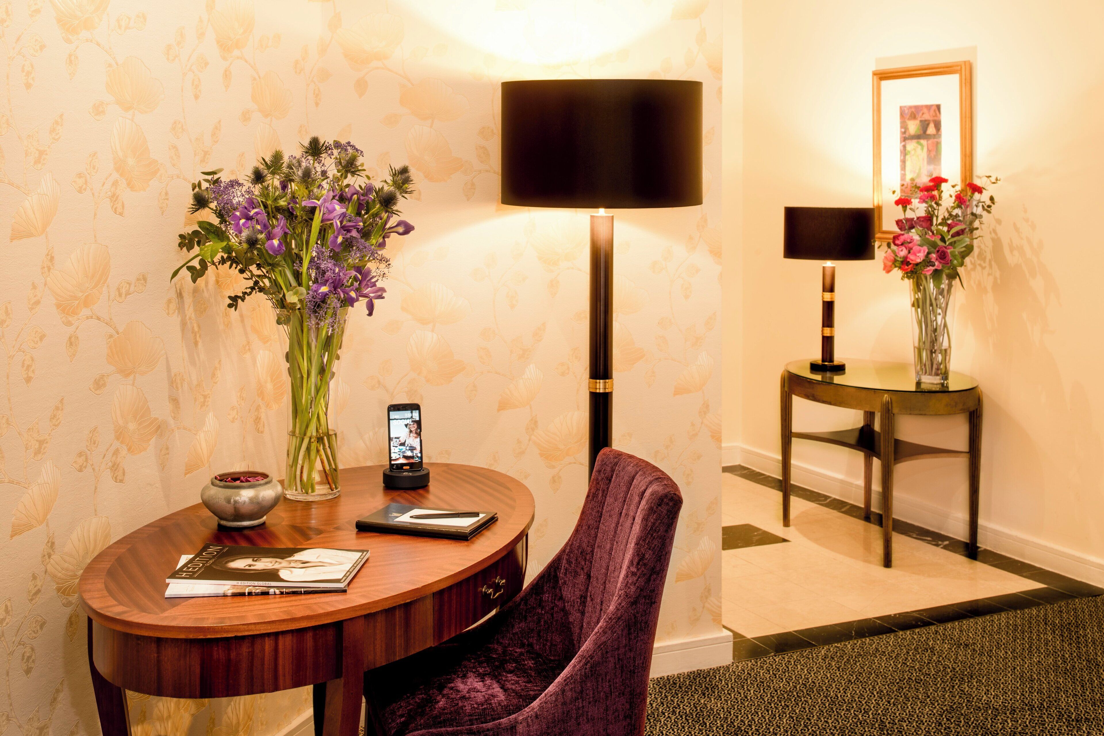 Een hotelkamer met een ronde houten tafel, een paarse stoel, een vaas met paarse bloemen, een lamp en een ingelijst beeld aan de muur.