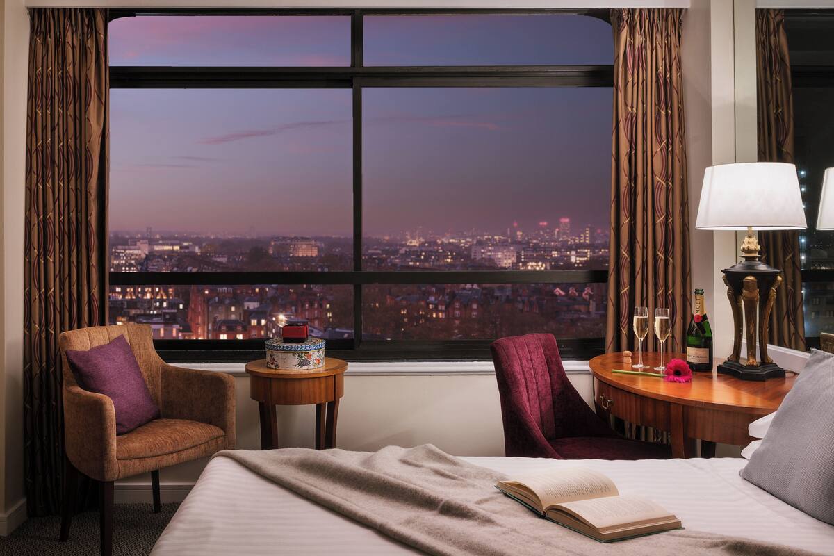 A hotel room with a large window offering a city view at dusk, a bed with a book, a chair, a small table with a bottle and glasses, and a lamp.
