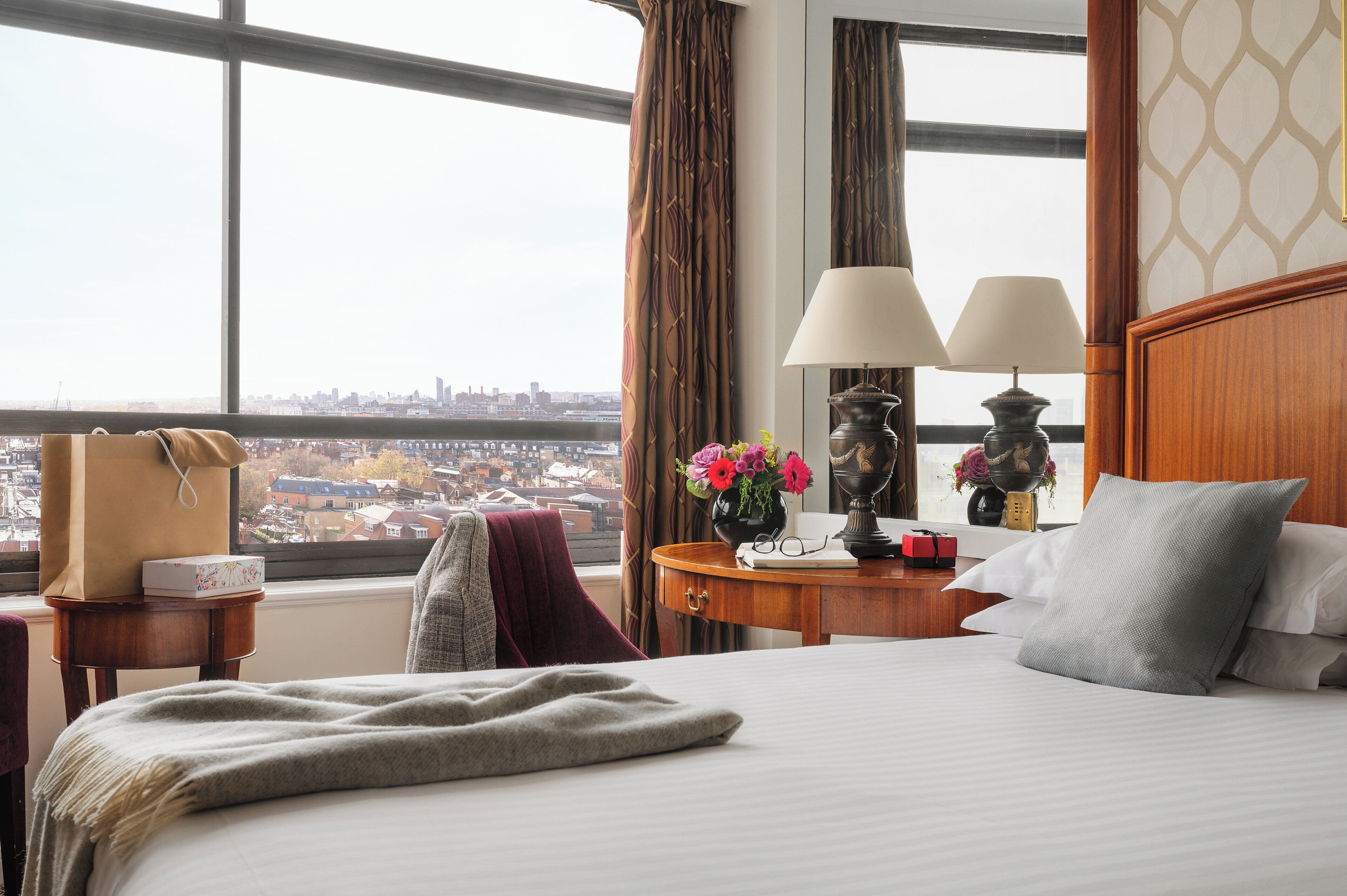 A hotel room with a large window, a bed with pillows, a bedside table with lamps, and a vase with flowers.