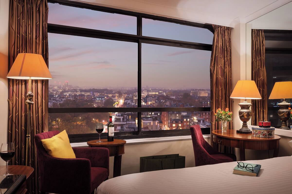 A hotel room with a large window offering a city view, a bed, a chair, a table with a lamp, and a vase with flowers.