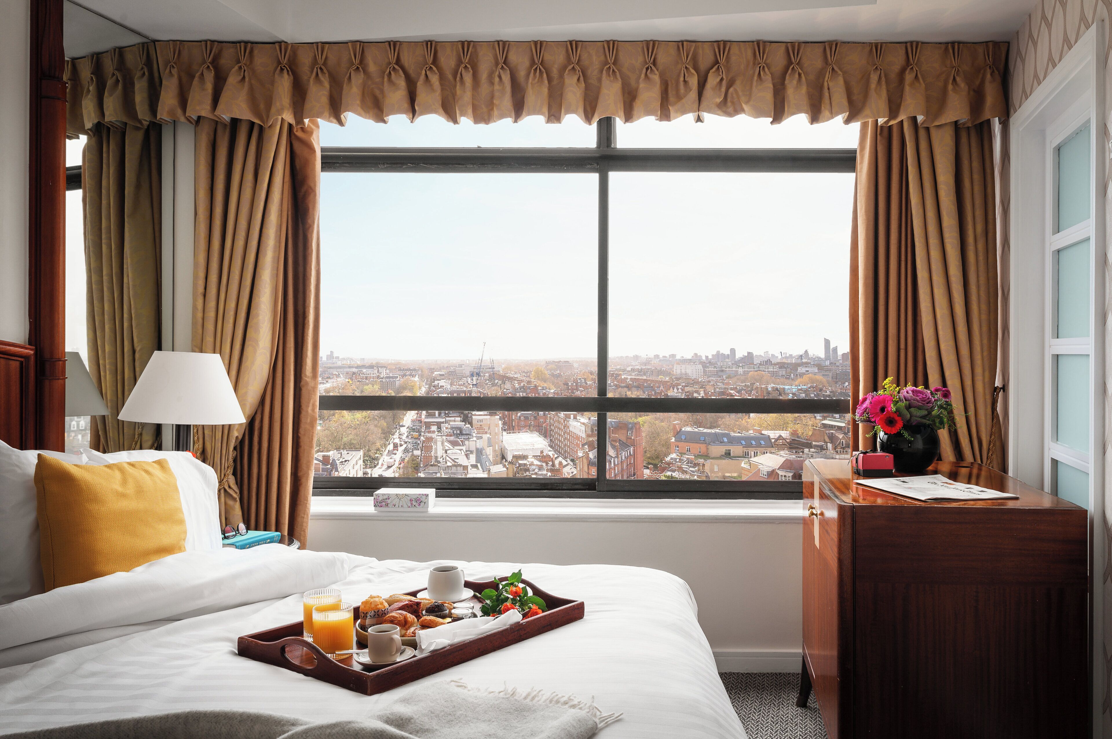 Ein Hotelzimmer mit großem Fenster, einem Bett mit Frühstücksbrett, einem Schreibtisch mit Blumenvase und einer Lampe.