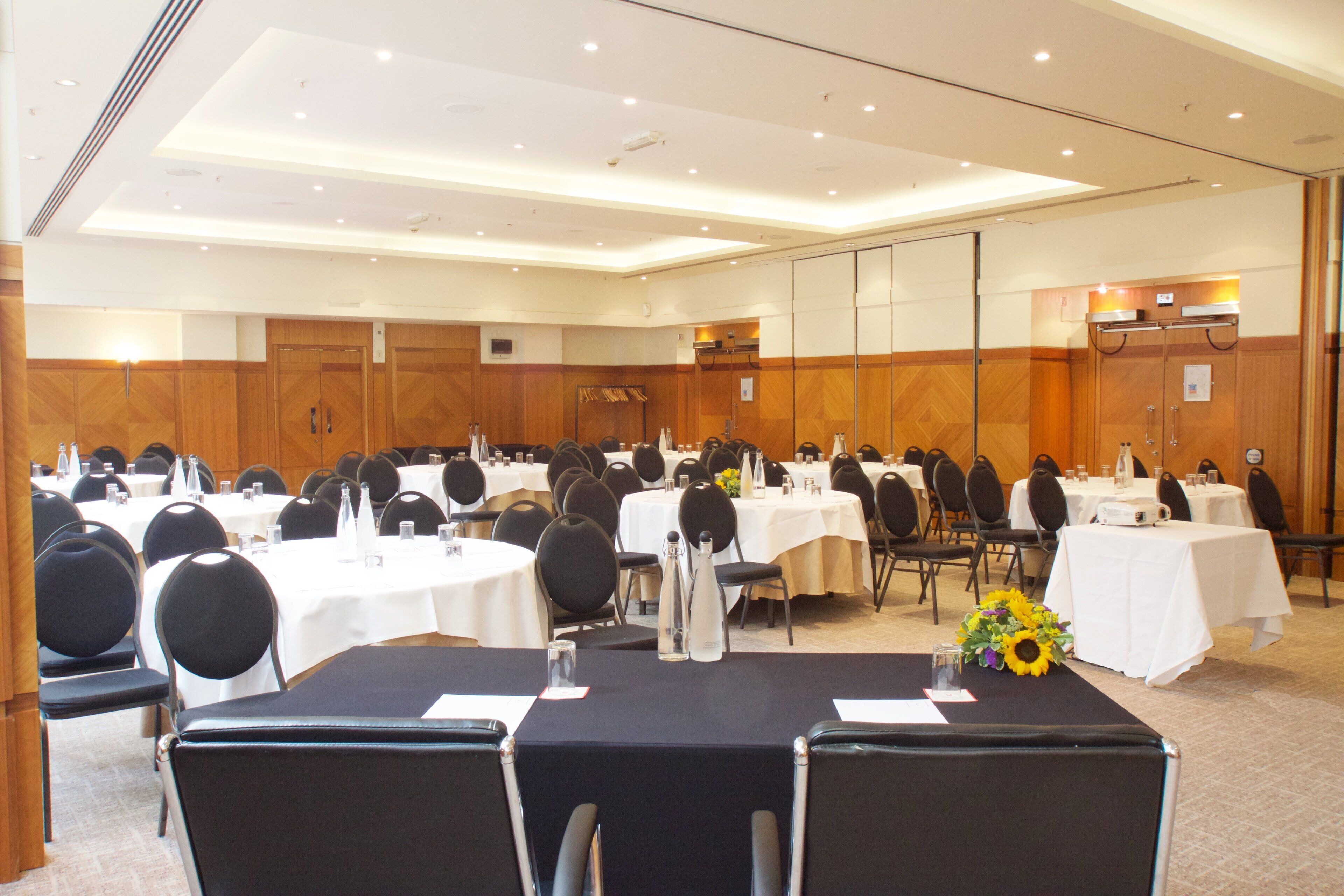 A conference room with multiple round tables, black chairs, and white tablecloths.