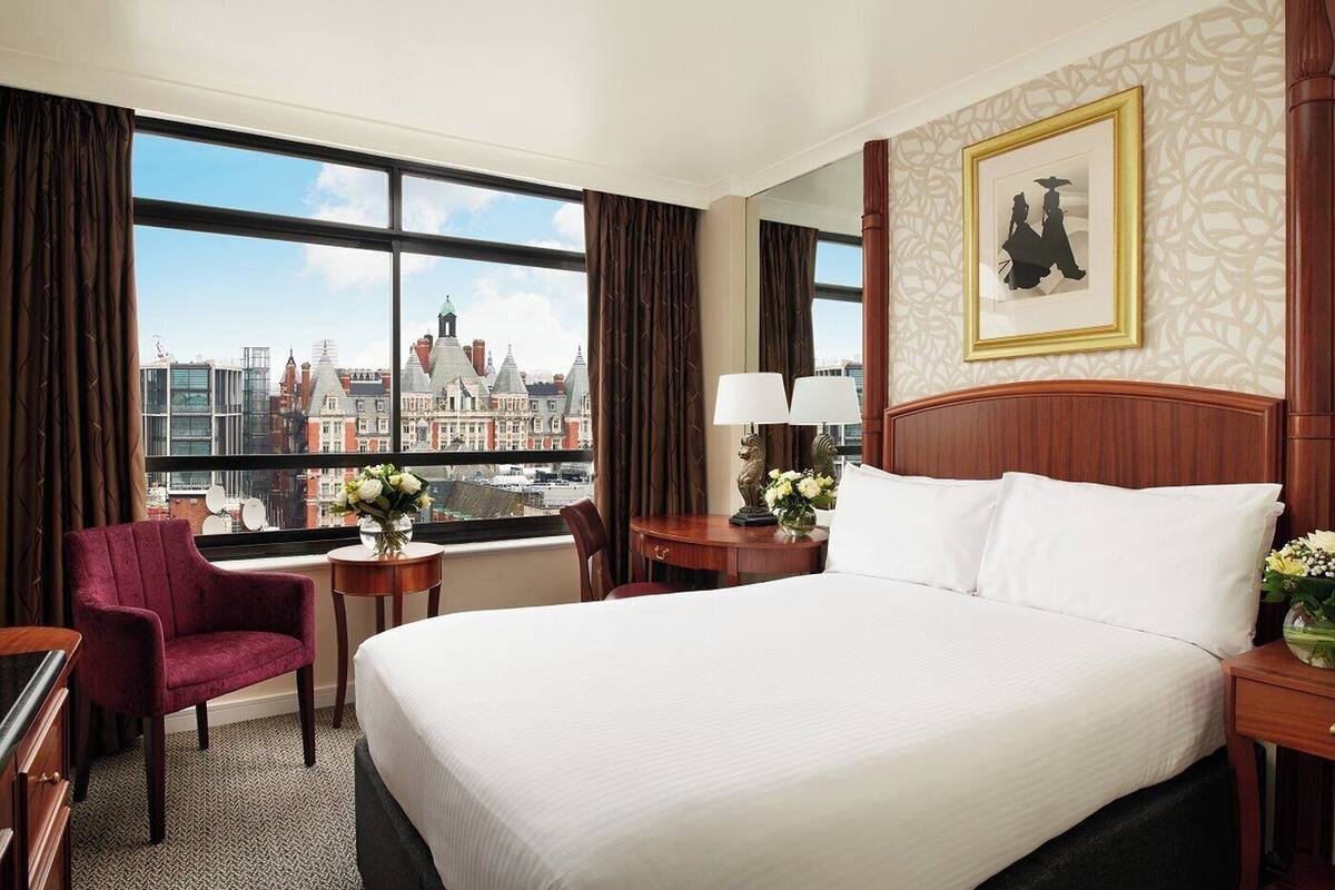 A hotel room with a large bed, a chair, a desk, and a view of a cityscape through large windows.