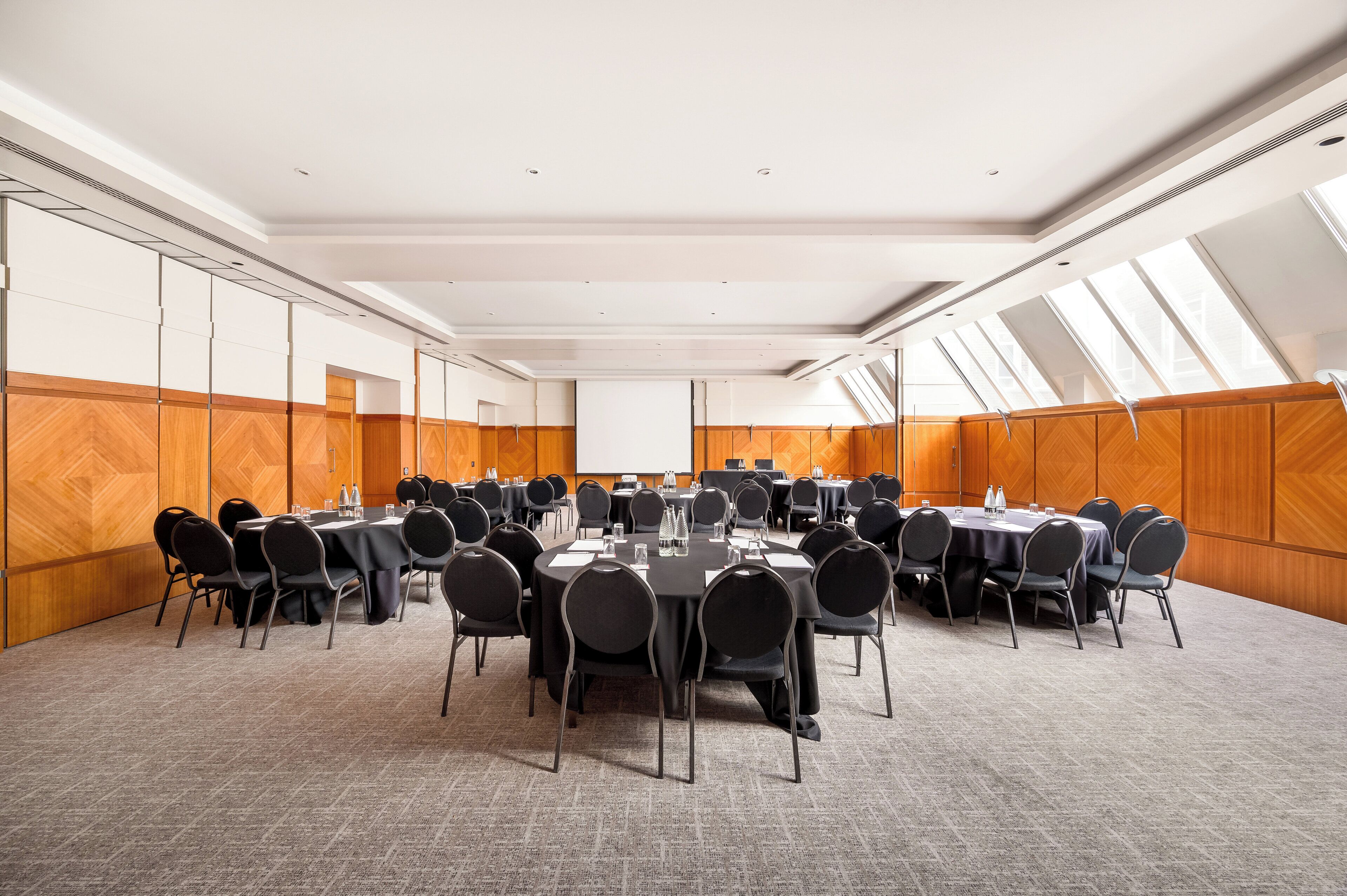 A conference room with multiple tables and chairs, large windows, and a carpeted floor.