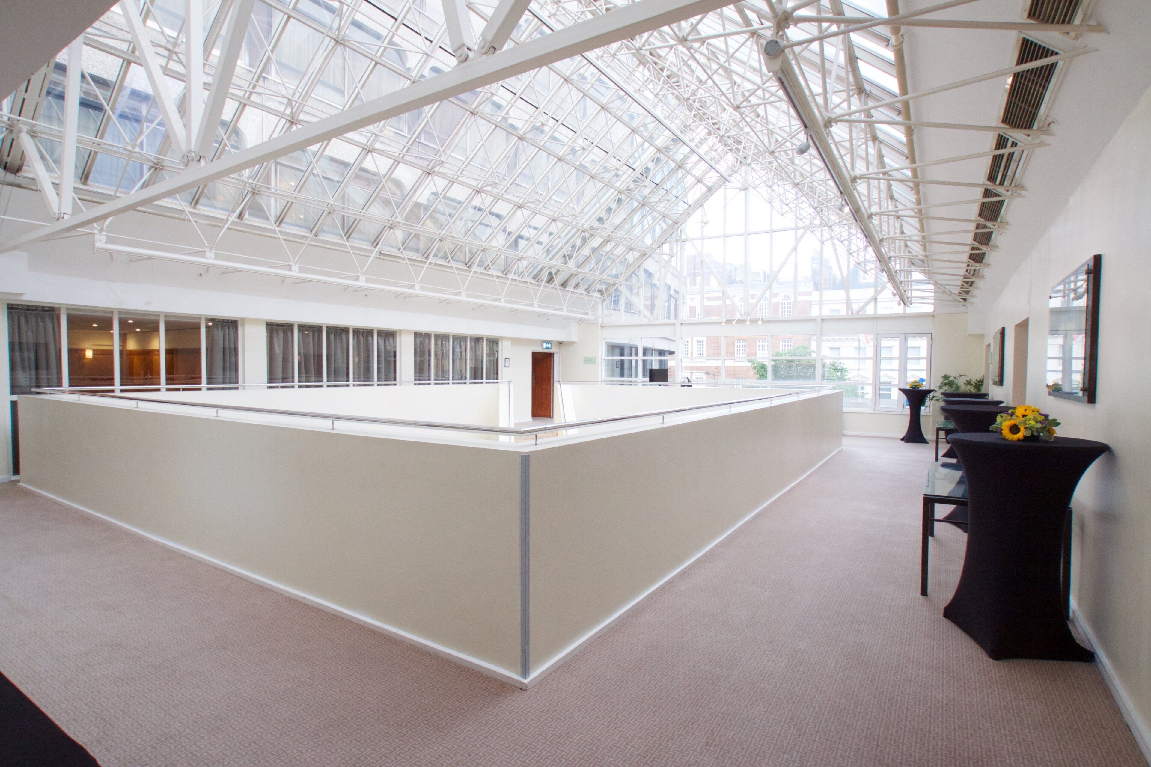 A spacious atrium with a high ceiling and large windows, featuring a central island with a wooden bench and potted plants.