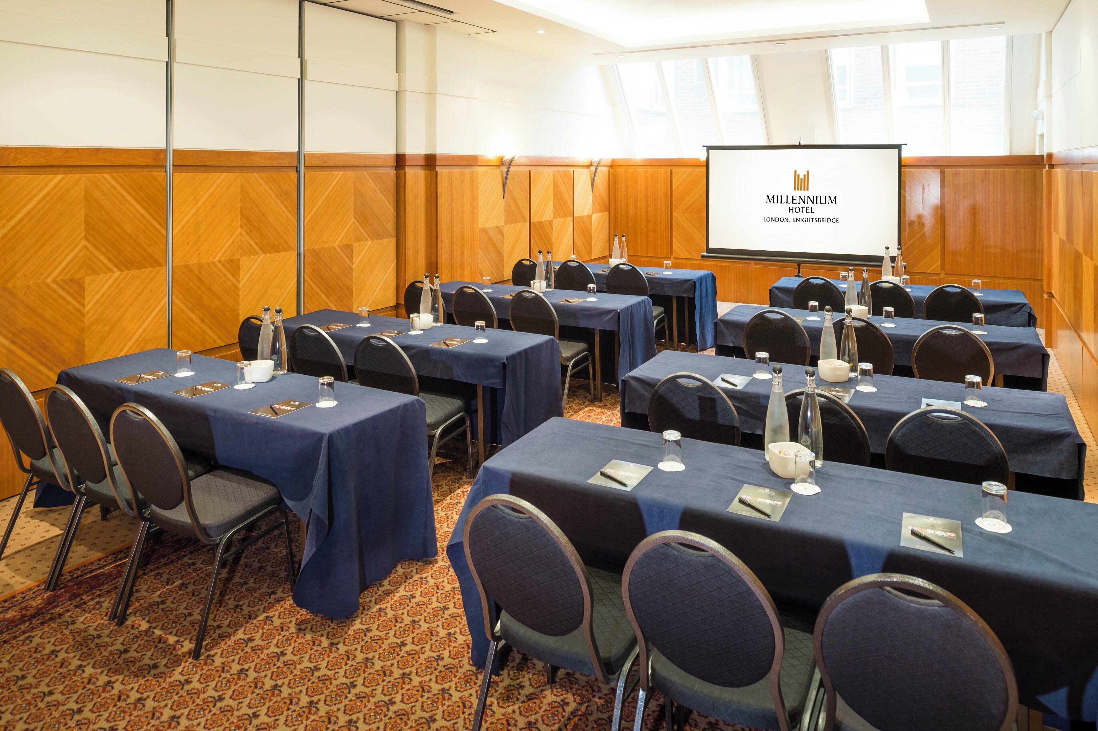 A conference room with tables set for meetings, a screen displaying the hotel name, and a patterned carpet.
