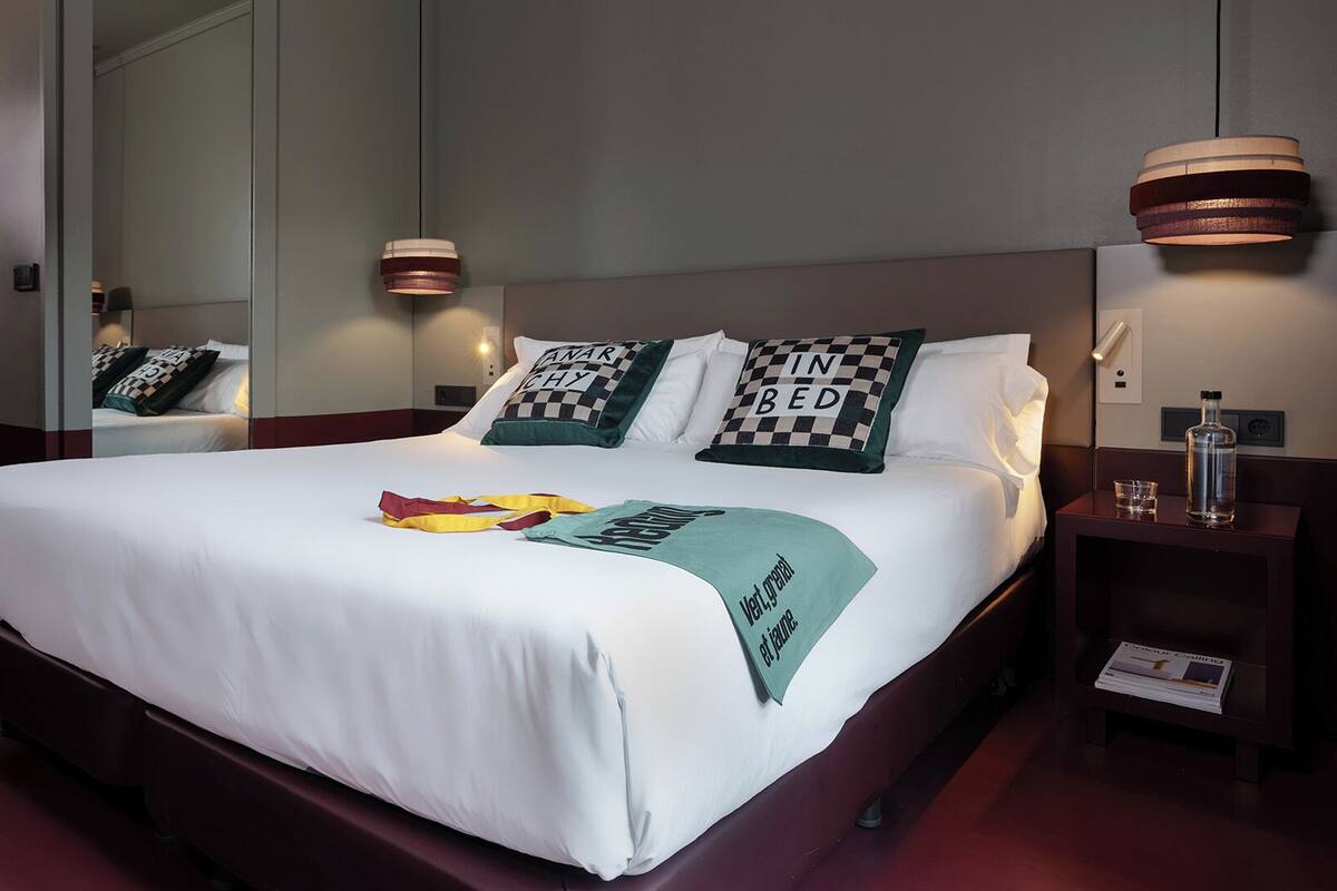 A hotel room with a large bed, two bedside lamps, a nightstand with a bottle and glass, and a mirror on the wall.