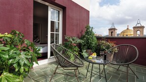 A terrace with two wicker chairs, a small table with a bowl of fruit and a bottle of wine, and a potted plant.
