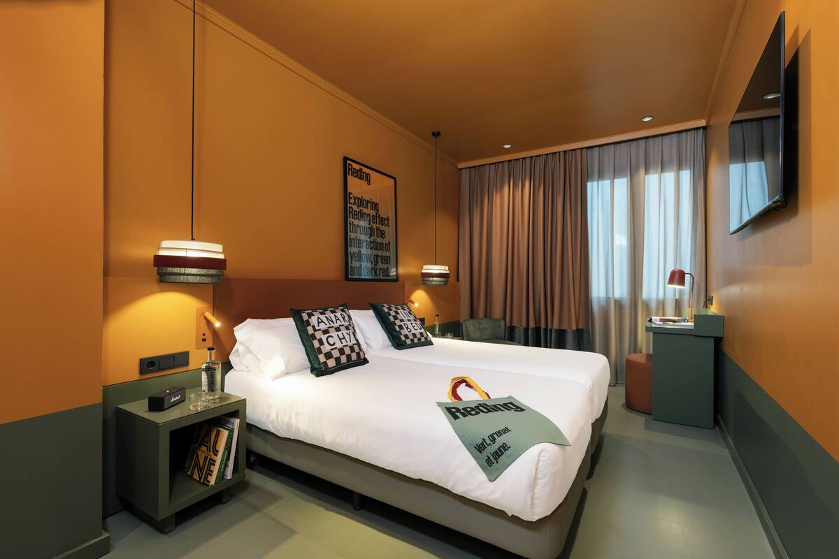 A hotel room with a large bed, two bedside tables, a nightstand, a desk, a chair, a TV mounted on the wall, and a window with curtains.