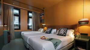 A hotel room with two beds, a nightstand with a lamp, a window with curtains, and a wall-mounted artwork.