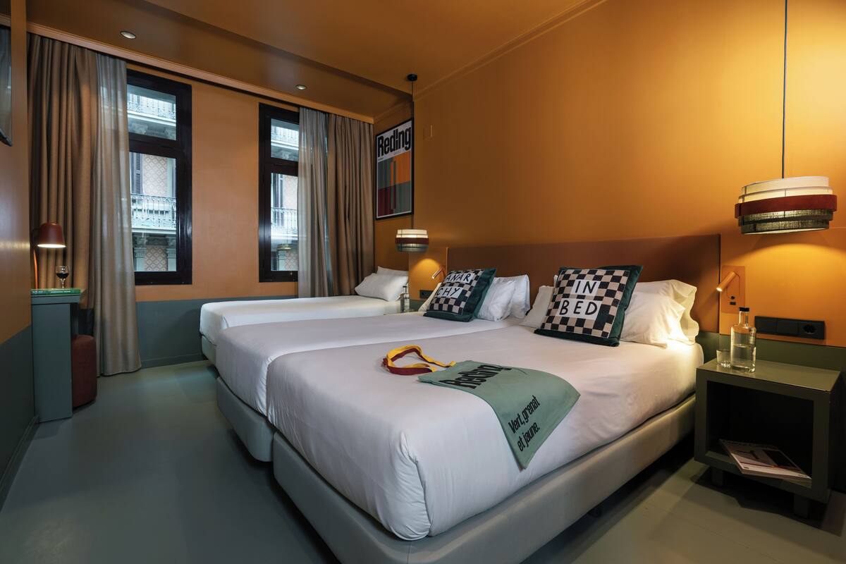 A hotel room with two beds, a nightstand with a lamp, a window with curtains, and a wall-mounted artwork.